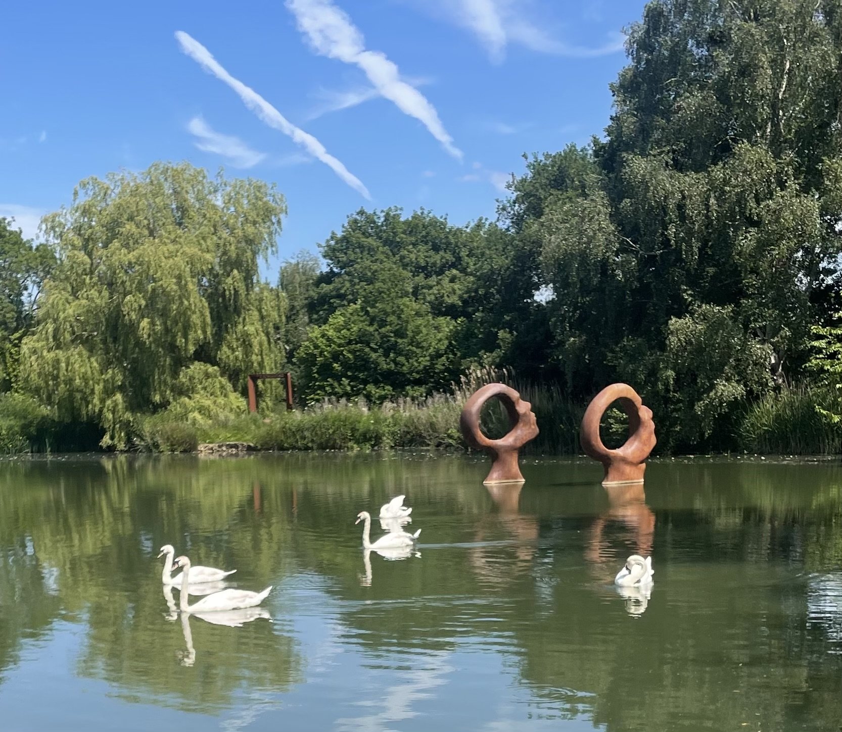 A pond with four swans swimming, lush trees and landscaped greenery in the background, sculptures on the water, and a blue sky with wispy clouds.
