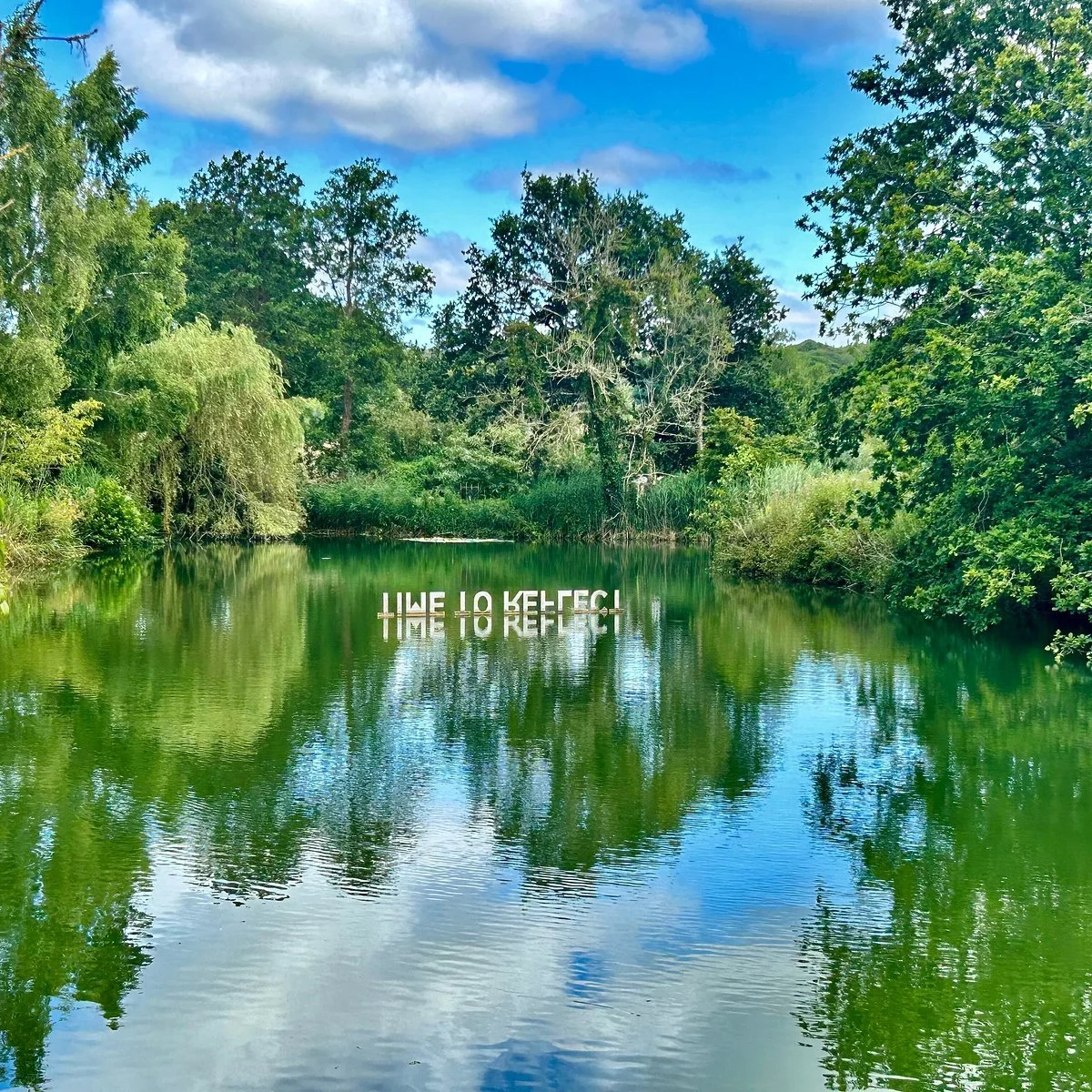 A calm river surrounded by lush green trees under a partly cloudy sky, with the phrase 'LIVE TO REFLECT' reflected on the water's surface.
