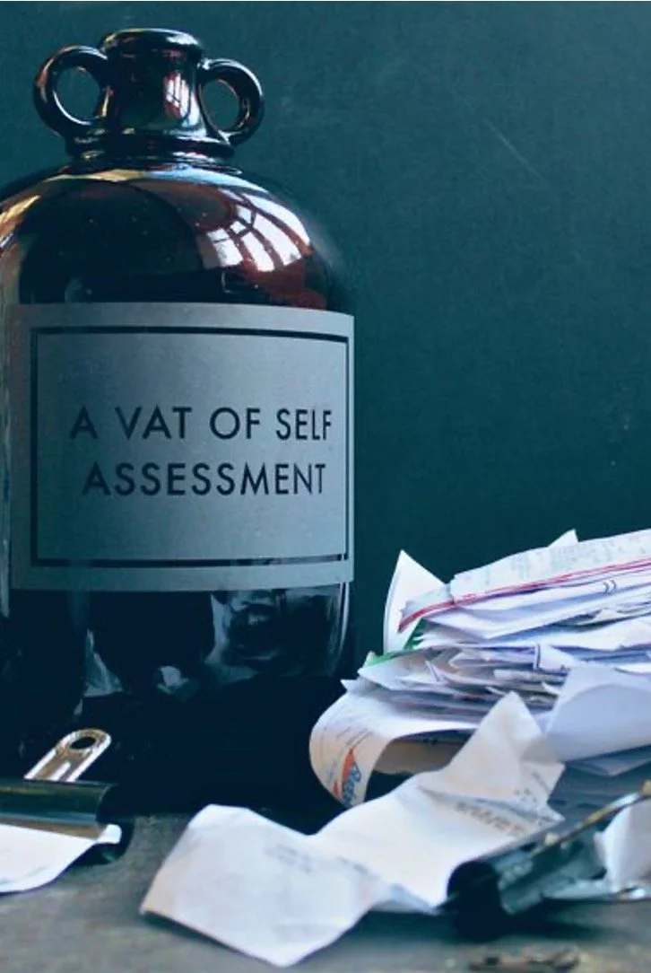 A dark-colored bottle labeled 'A VAT OF SELF ASSESSMENT' is placed next to a cluttered pile of papers and documents on a desk.