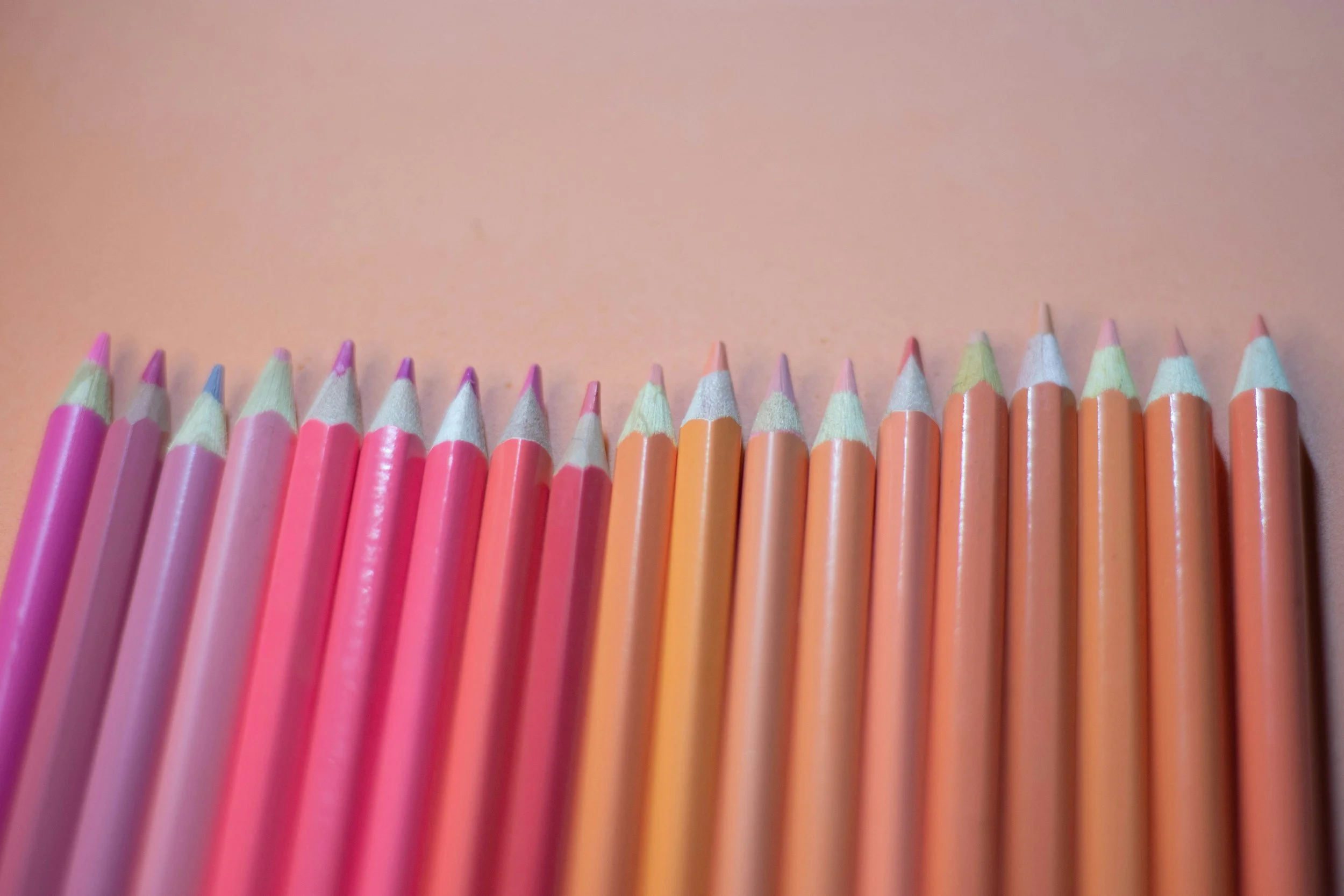 A row of colorful pencils in shades of pink, orange, and purple arranged on a pink background.