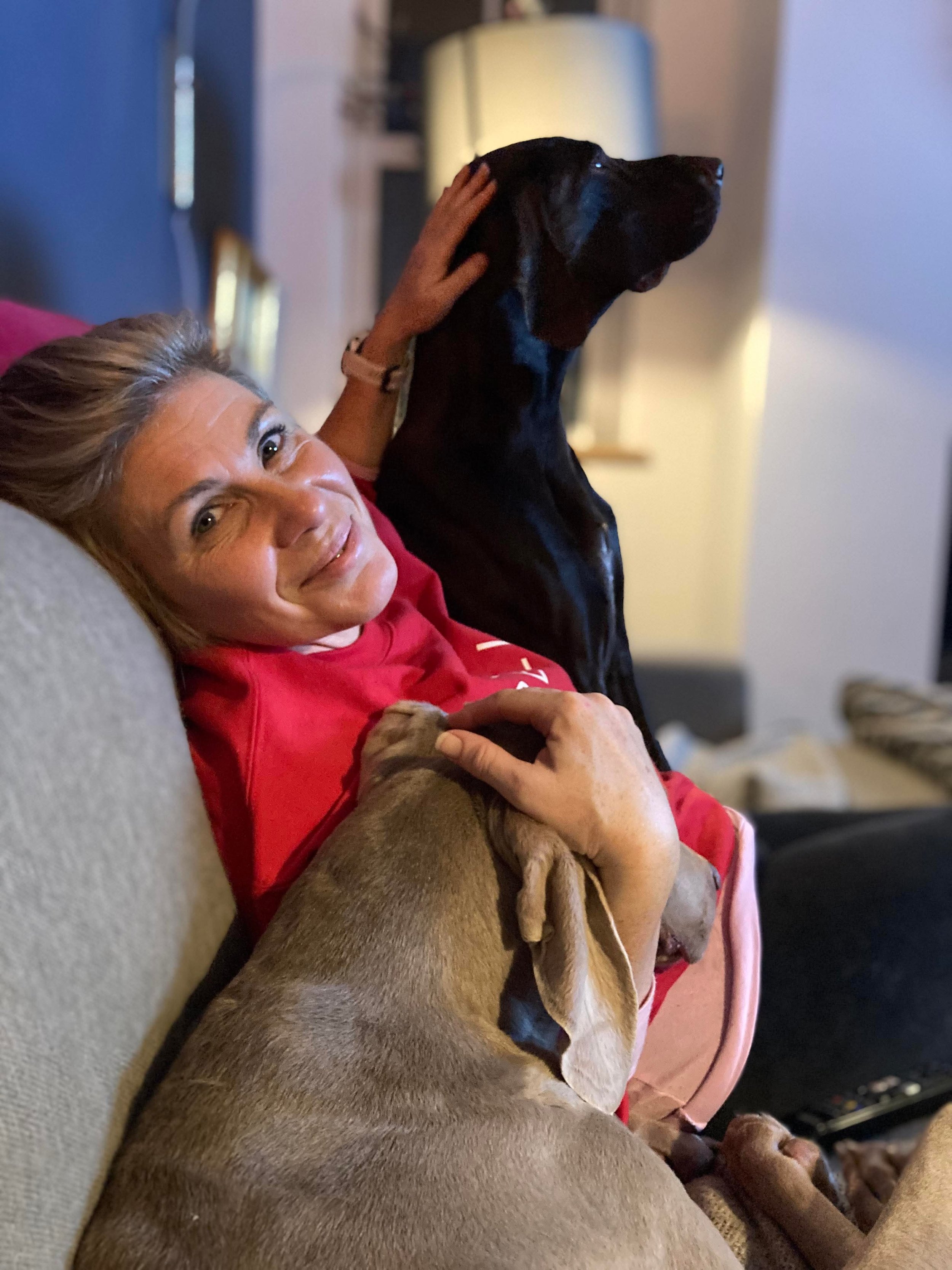 Danielle Brooks, founder of Brooksmiths, on a couch, smiling, cuddling two her puppies: a large silver dog (weimaraner) resting its head on her shoulder and a large brown GSP dog with her hand on the back of its head.