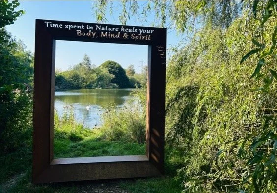 A large rectangular mirror outdoors on grass next to trees and bushes, reflecting a river, trees, and blue sky with clouds, with a quote on top that reads, "Time spent in Nature heals your Body, Mind & Spirit."
