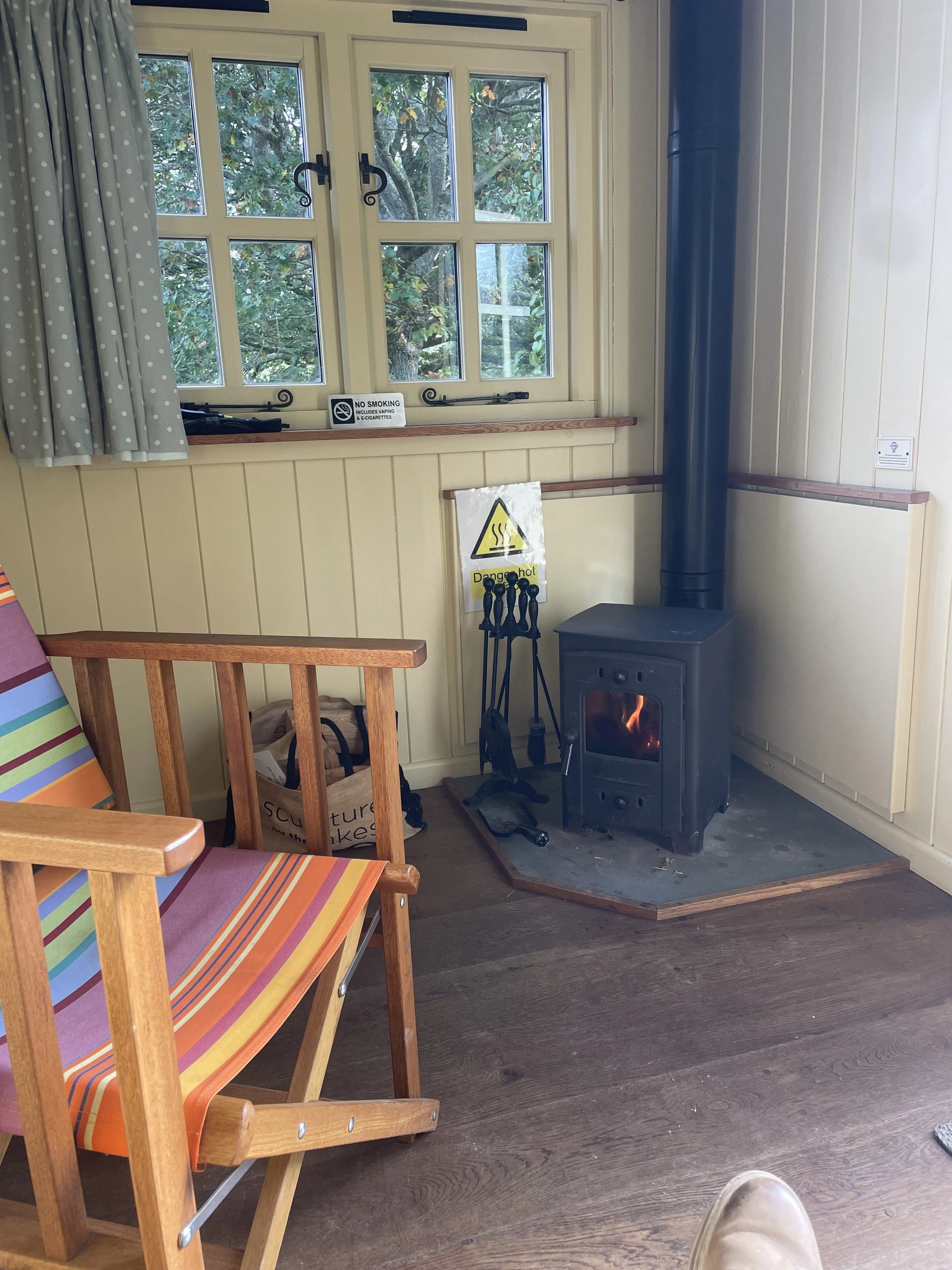 A cozy indoor corner featuring a wood stove with a chimney pipe, a small wooden bench with a colorful striped fabric seat, a paper bag on the floor, and a window with pale green wooden frames and blue polka-dot curtains. The window shows green foliage outside. There are no smoking or hot surface signs on the wall near the stove.