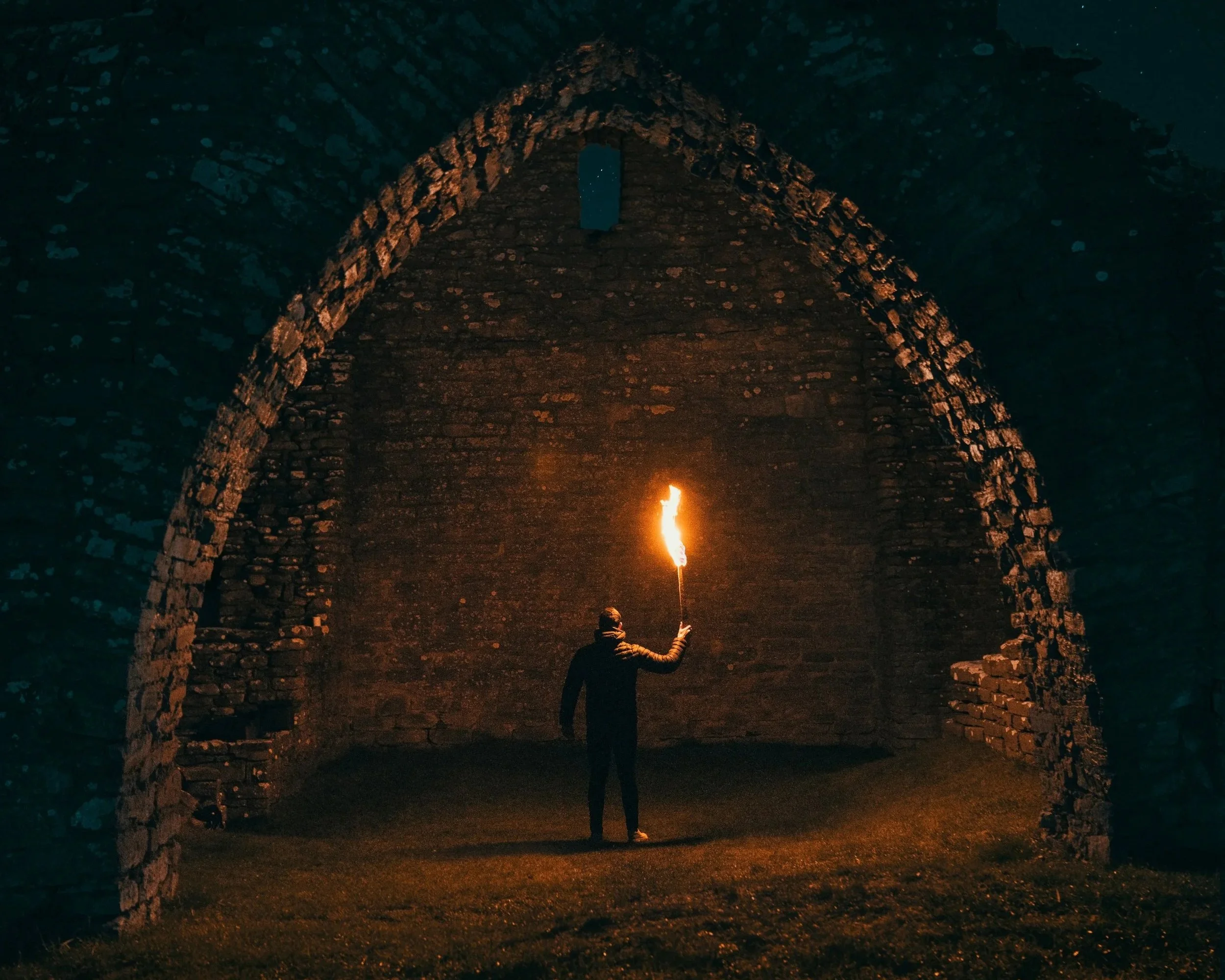 A person holding a lit torch inside an ancient stone archway at night.
