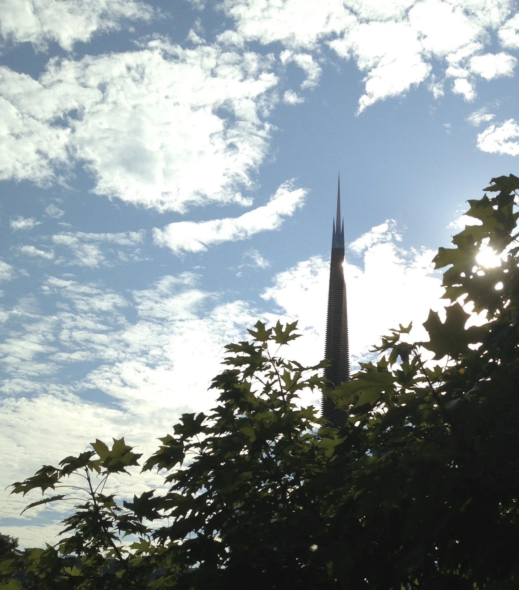 Spear statue behind maple tree pointing to wispy clouded blue sky