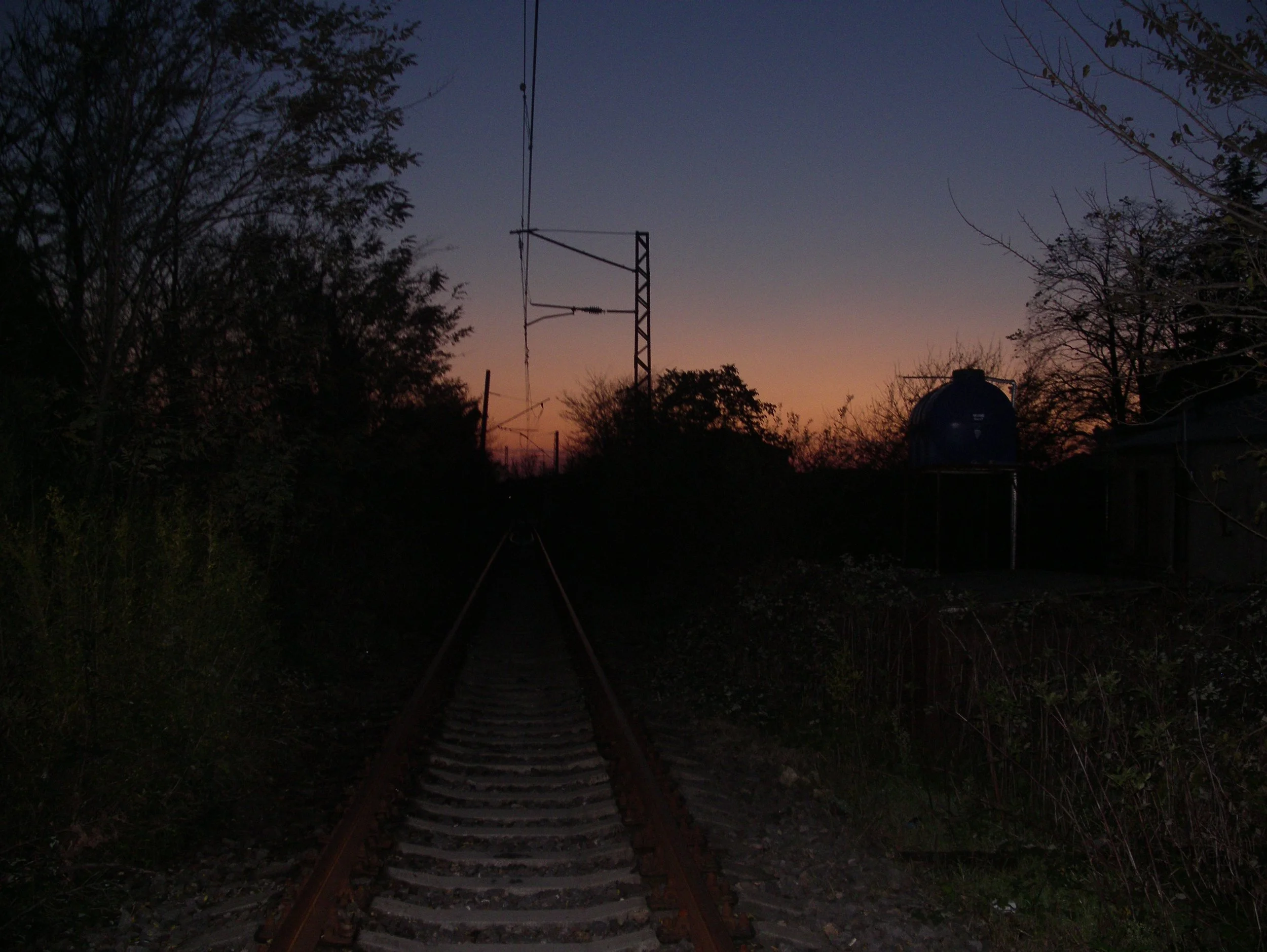 A dark railway track during dusk with trees and a water tank on the right, and a power line and pole in the background.