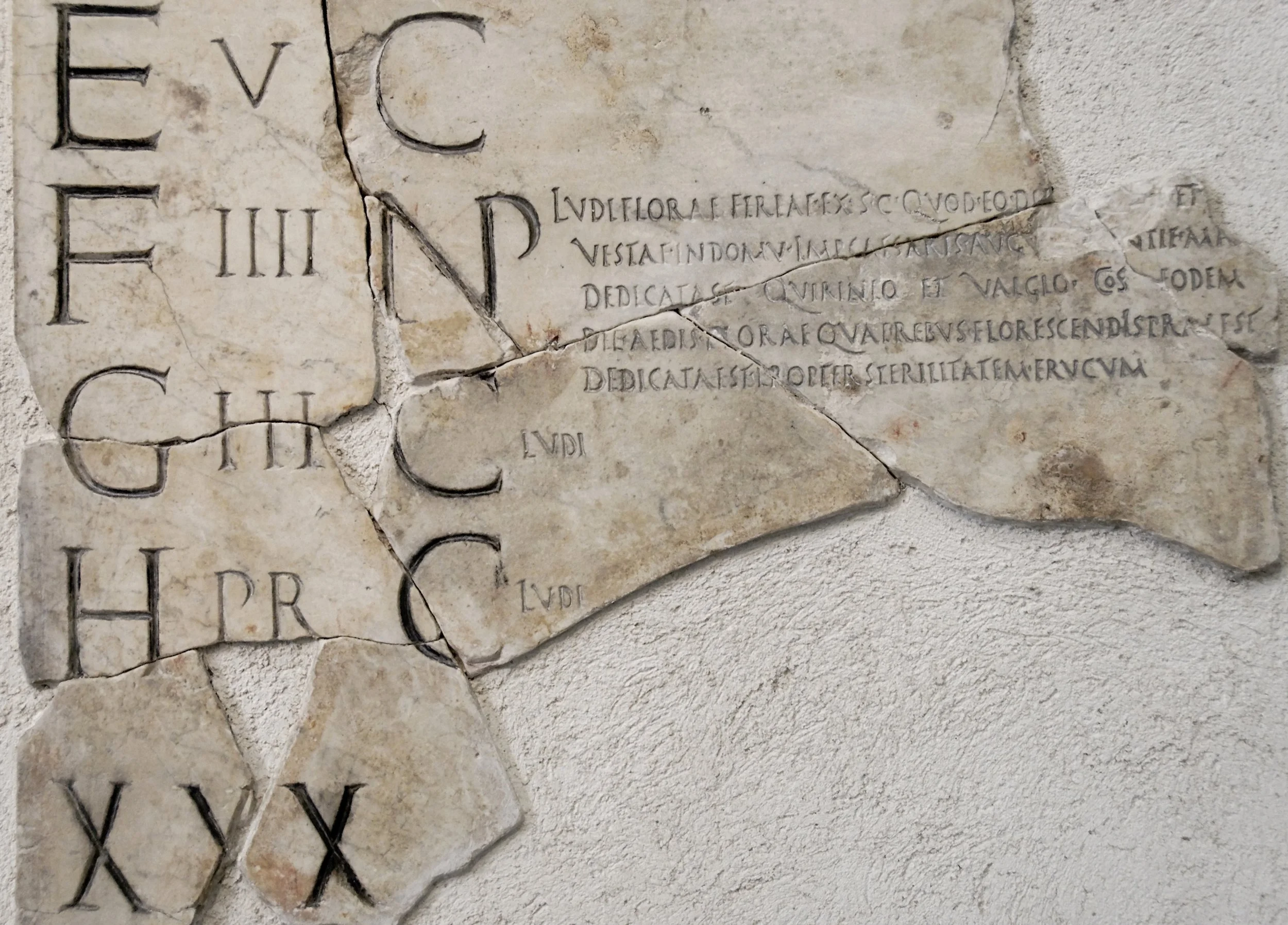 Broken ancient stone inscription with Latin script, partially damaged and cracked, mounted on a wall.