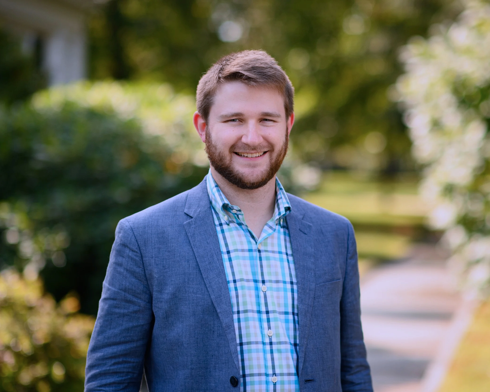 A young man with a beard and short brown hair, wearing a blue plaid shirt and a gray blazer, standing outdoors in a park during daytime, smiling at the camera.