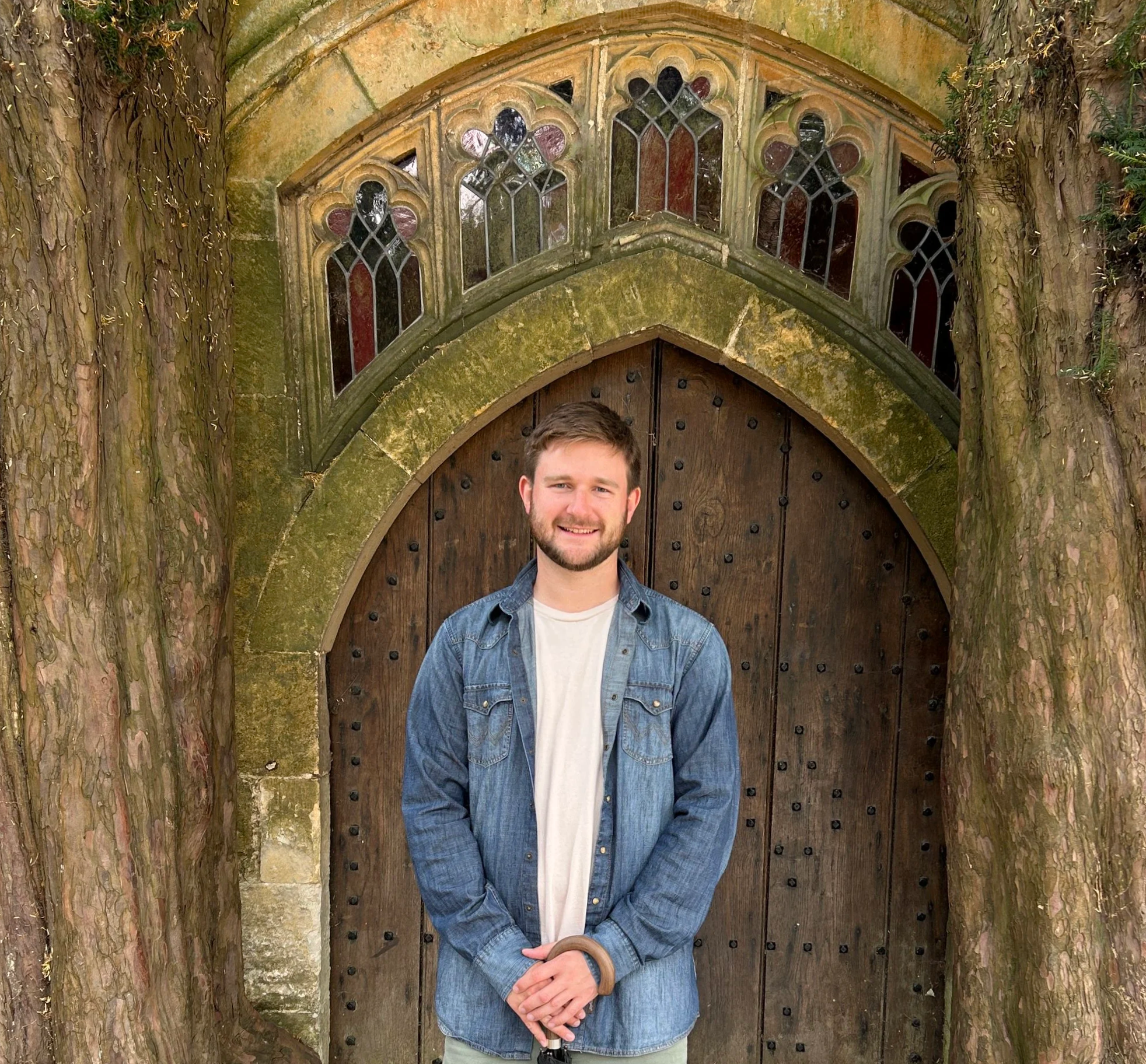 A young man with a beard smiling and standing in front of a large wooden door set into a stone archway, framed by tree trunks, with stained glass windows above.
