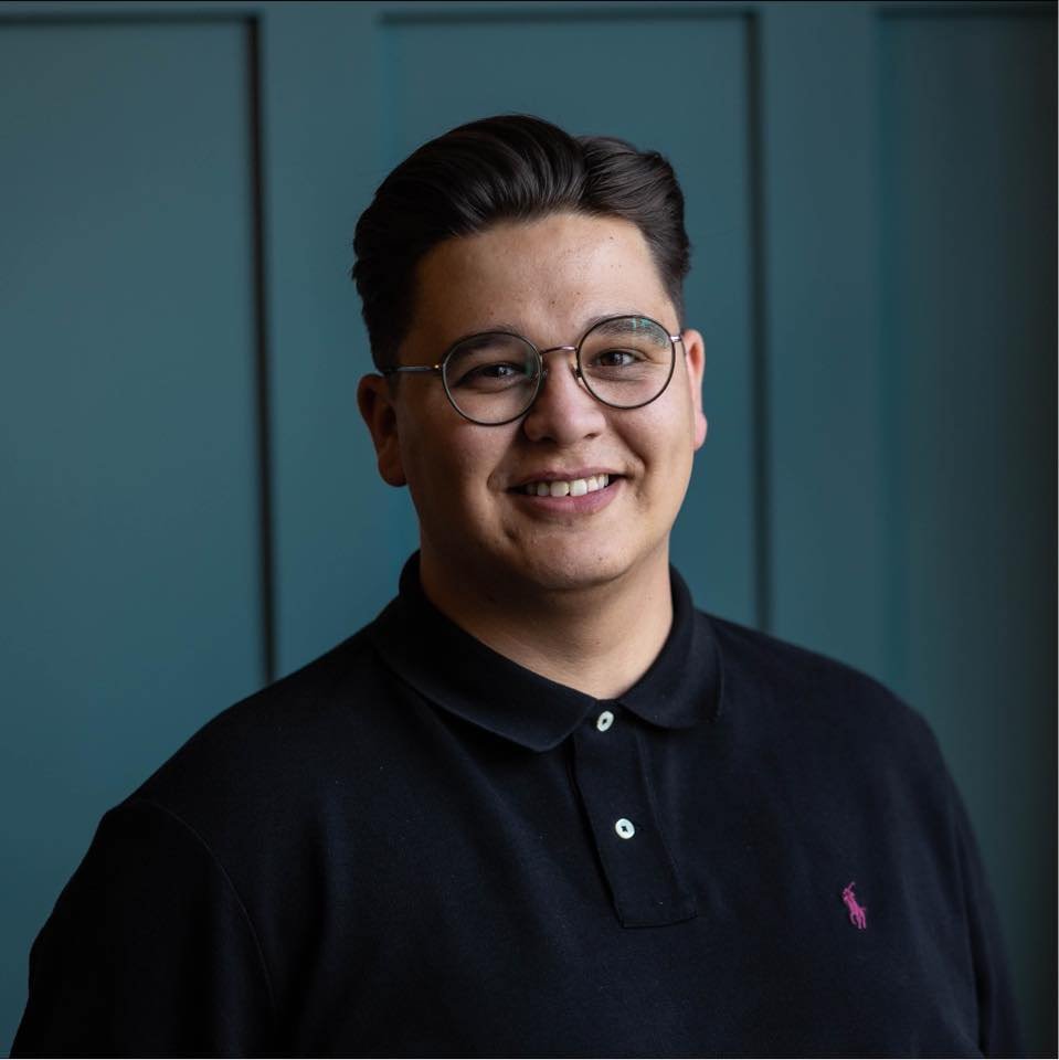 A young man with dark hair, glasses, and a black polo shirt with a pink logo, standing in front of a plain teal wall and smiling at the camera.