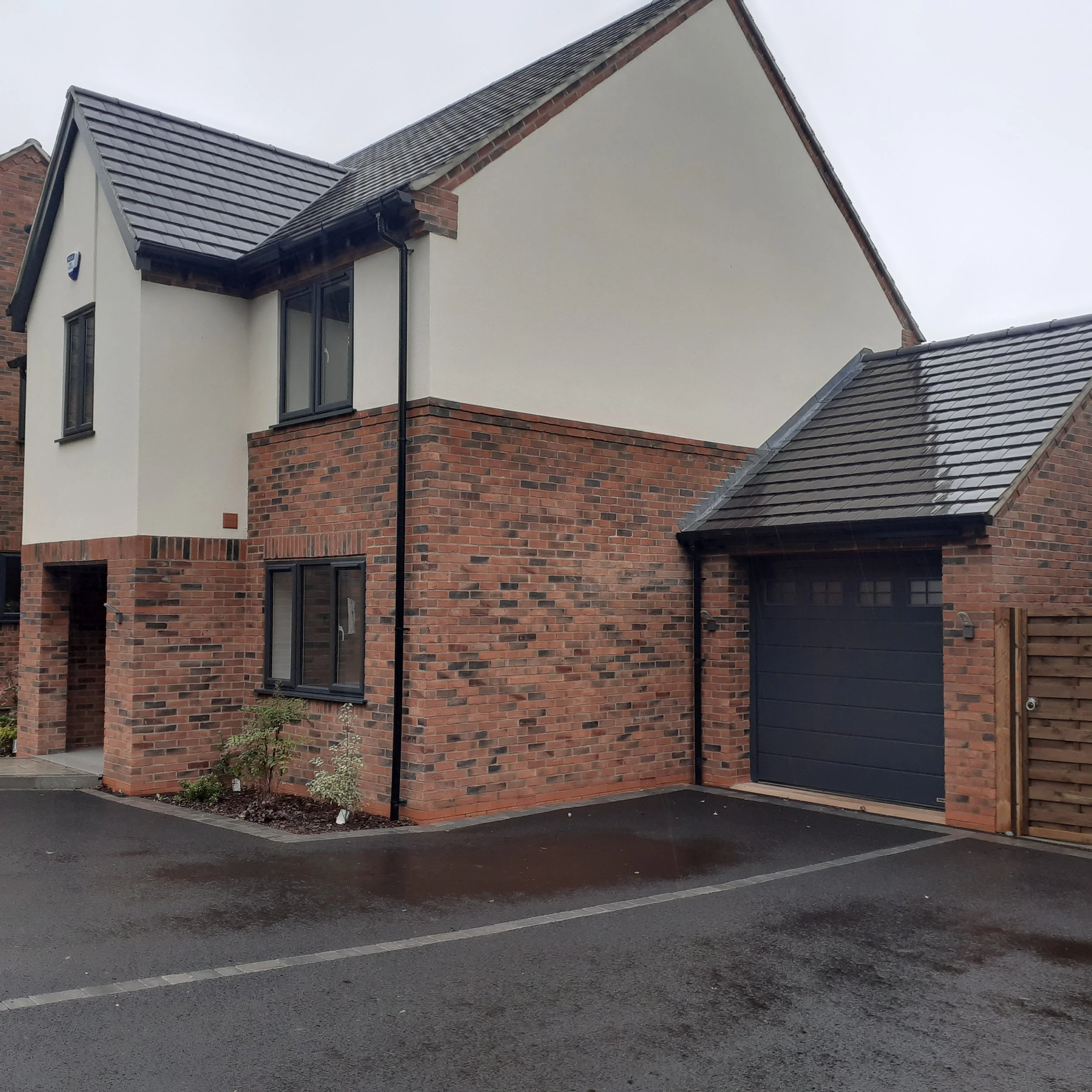 Modern two-story house with brick and white render exterior, black window frames, and a black garage door, situated on a wet driveway under overcast skies.