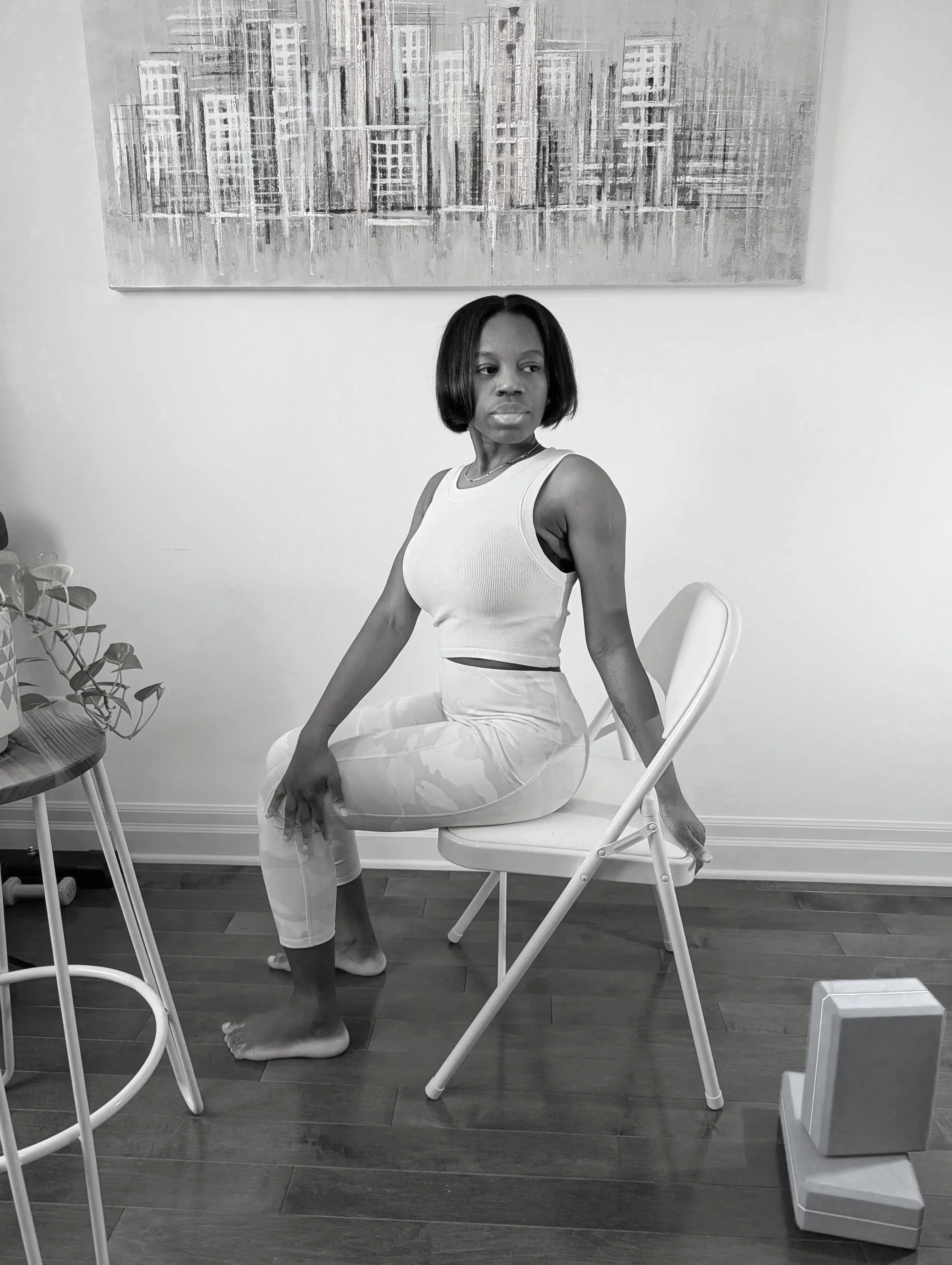 Une femme assise sur une chaise, regardant de côté, dans une pièce avec une peinture abstraite en arrière-plan, en noir et blanc.