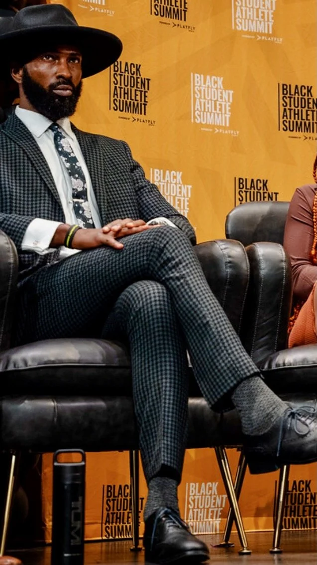A man in a black hat, checkered suit, and tie sitting on a black leather couch at the Black Student Athlete Summit event.