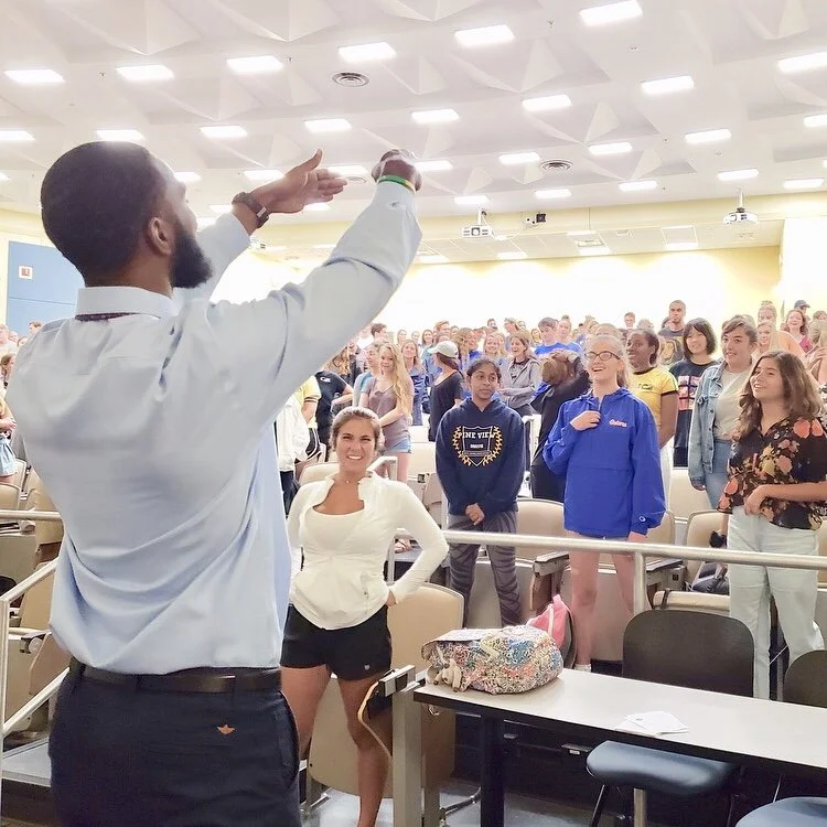 A man with a beard and short hair in a white shirt addressing a large group of diverse students in an auditorium. The students are standing, listening attentively, some smiling and some with hands on their hips. The auditorium has bright ceiling ligh