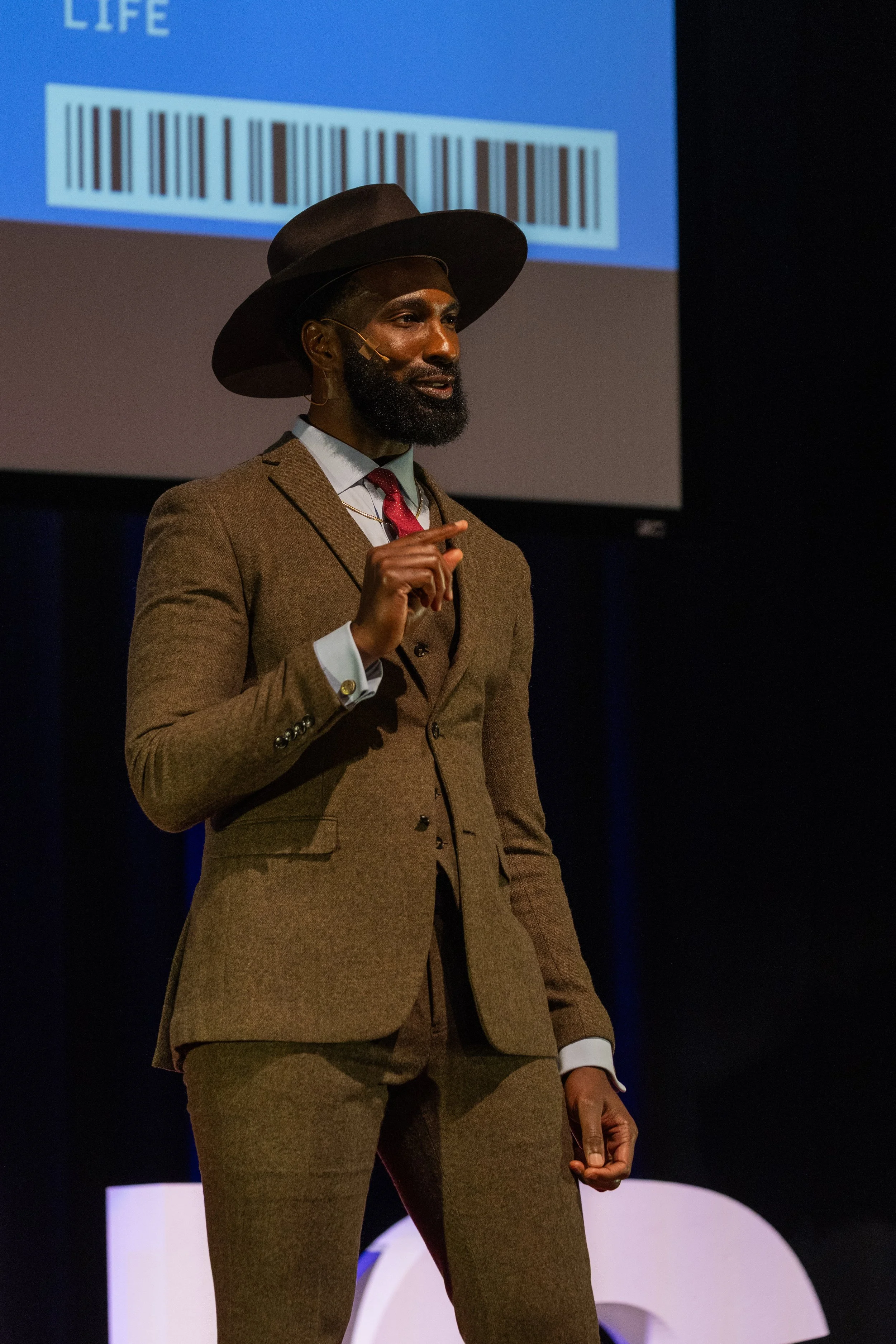 A man dressed in a brown suit, white shirt, and red tie, wearing a wide-brimmed hat, standing on stage with a black background and a large screen behind him displaying a barcode and the word 'LIFE'.