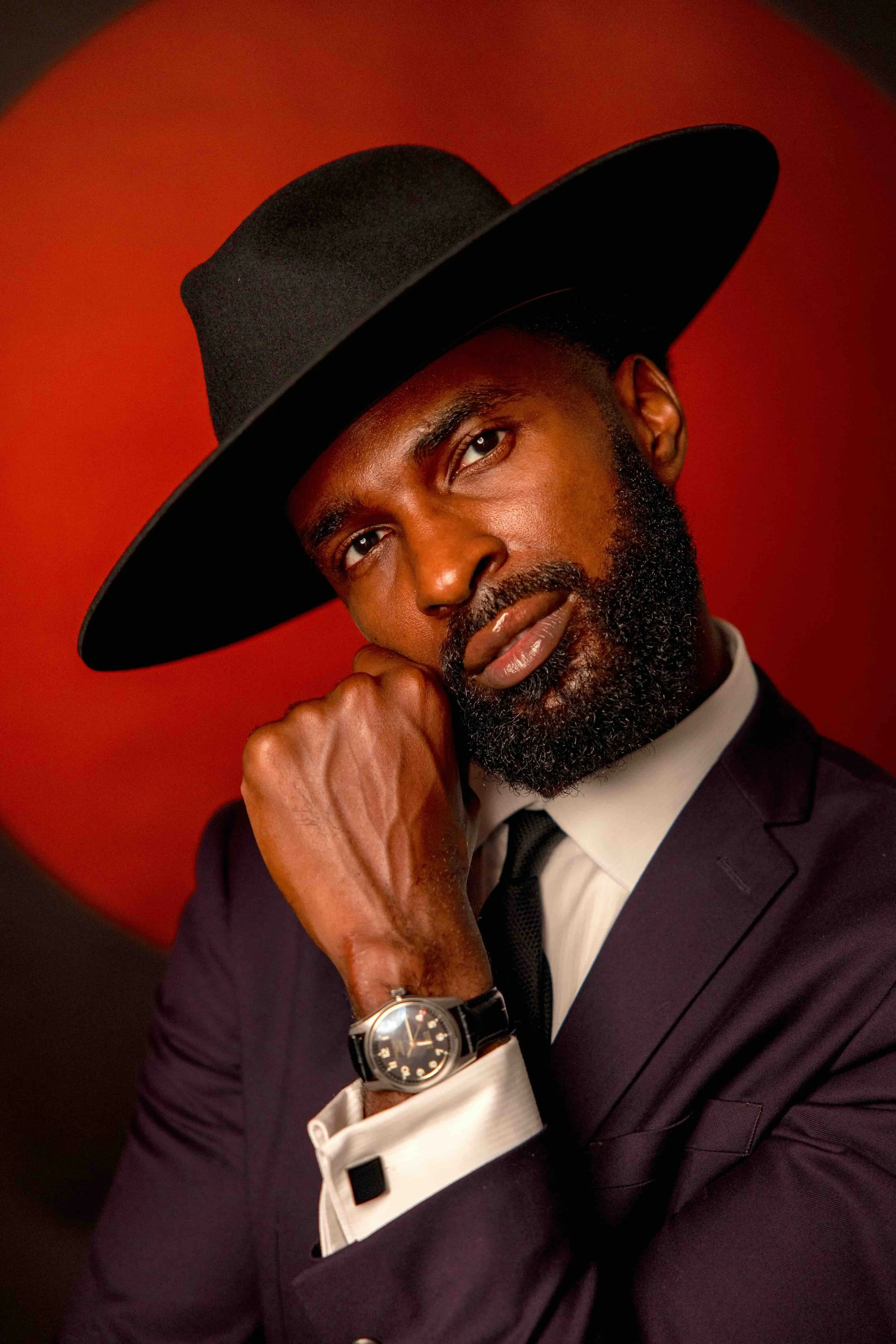 A man wearing a black wide-brimmed hat, black tuxedo, white shirt, and black tie, with a beard and mustache, resting his chin on his fist, against a red background.