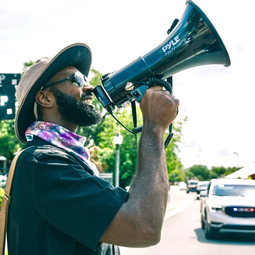 A man wearing sunglasses and a wide-brimmed hat is holding a megaphone and speaking into it outdoors during daytime, with cars and trees in the background.