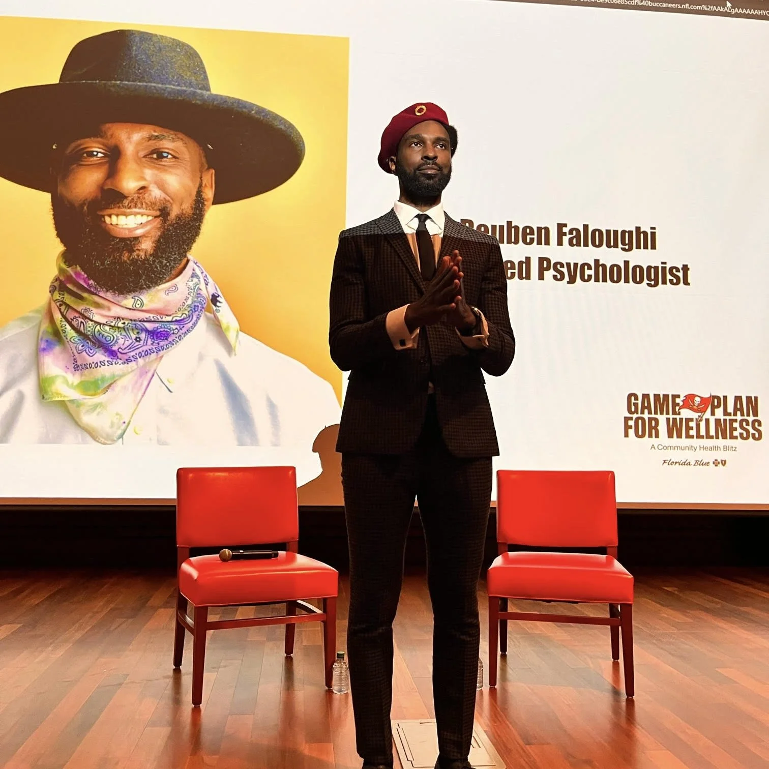 A man in a dark suit with a burgundy beret stands with hands together in front of a large poster that features a smiling man with a beard wearing a large hat and colorful scarf. The poster has text mentioning Douben Faloughi, a psychotherapist, and a