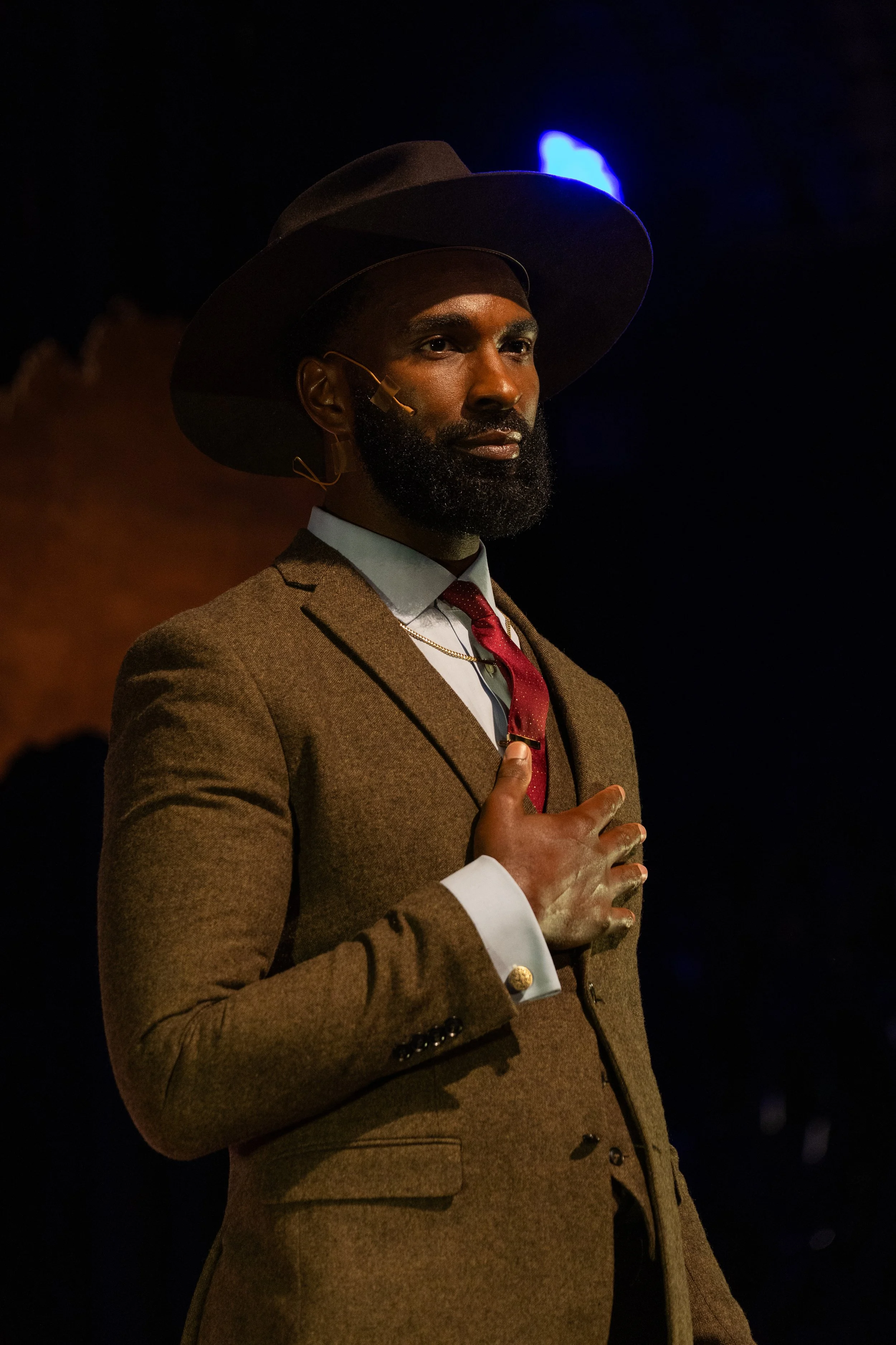 A man with dark skin, a full beard, dressed in a brown suit, white shirt, and red tie, wearing a wide-brimmed dark hat. He has a microphone attached to his face and his hand over his heart, standing against a dark background with blue and orange lights.