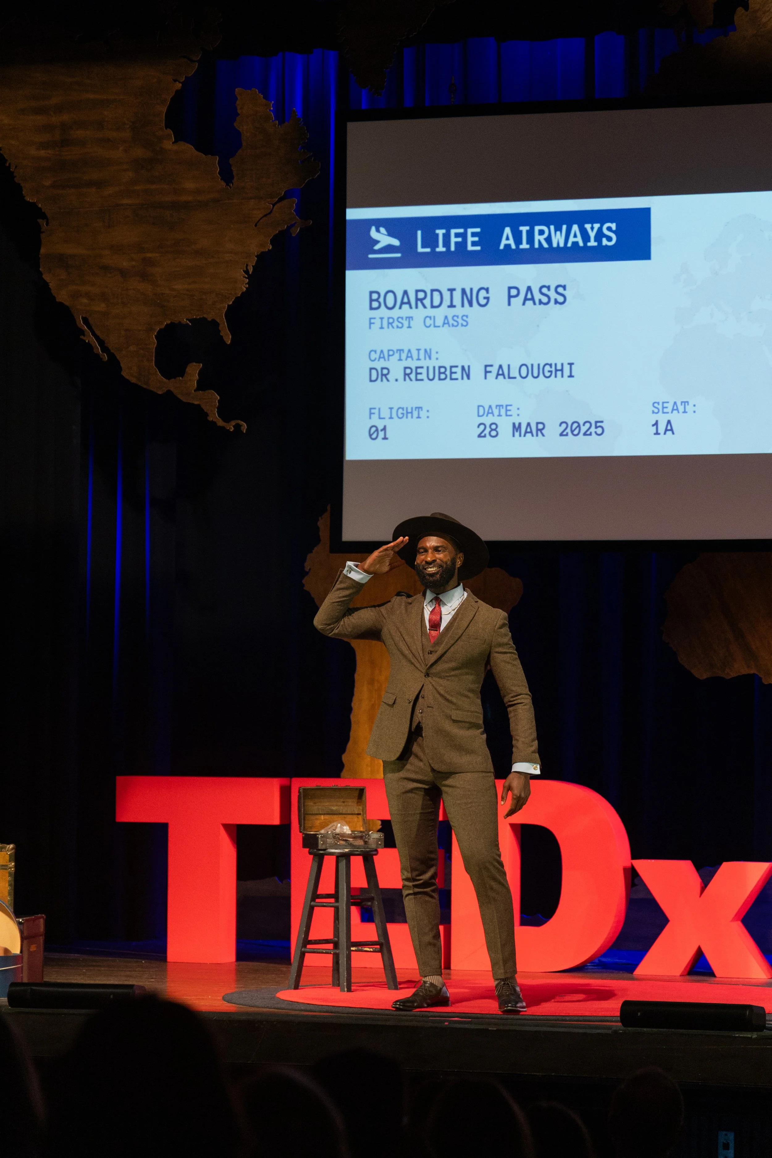 A man in a brown suit, black hat, and red tie on stage giving a TED Talk. There is a large red TED logo behind him, a stool, and a small table with props. A large screen displays a boarding pass for Life Airways with details including flight number 0