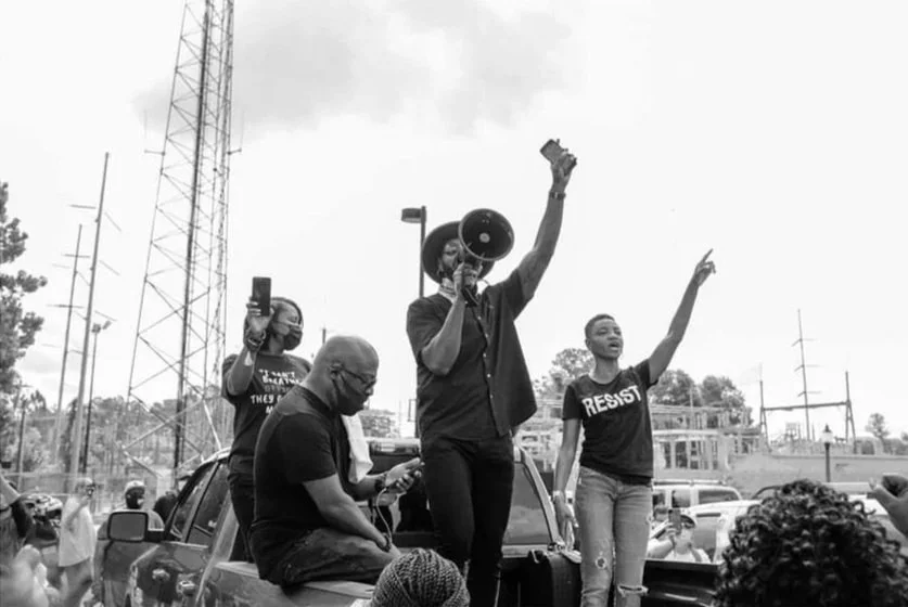 People participating in a protest or rally outdoors, with one person speaking into a megaphone, another holding a phone, and others raising their hands, some wearing t-shirts with messages like 'RESIST'.