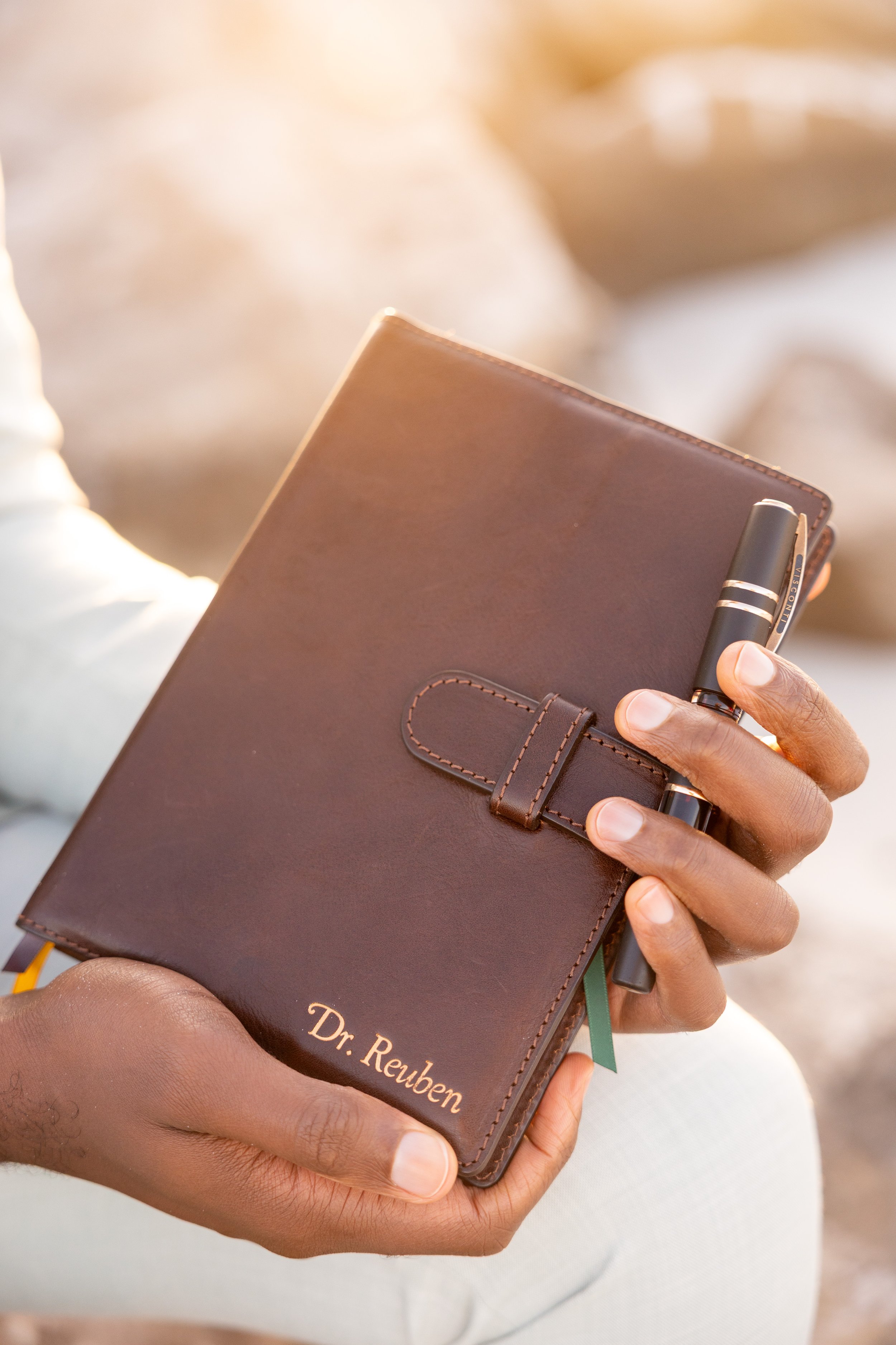 A person holding a brown leather briefcase with the name 'Dr. Reuben' engraved on it, along with a black pen clipped to the briefcase strap.