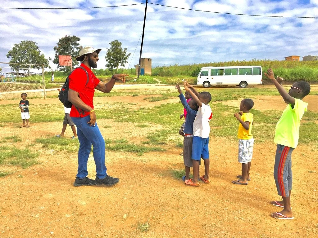 A man with a backpack and hat is teaching a group of children outdoors on a dirt field, with children raising their hands to answer or ask questions.