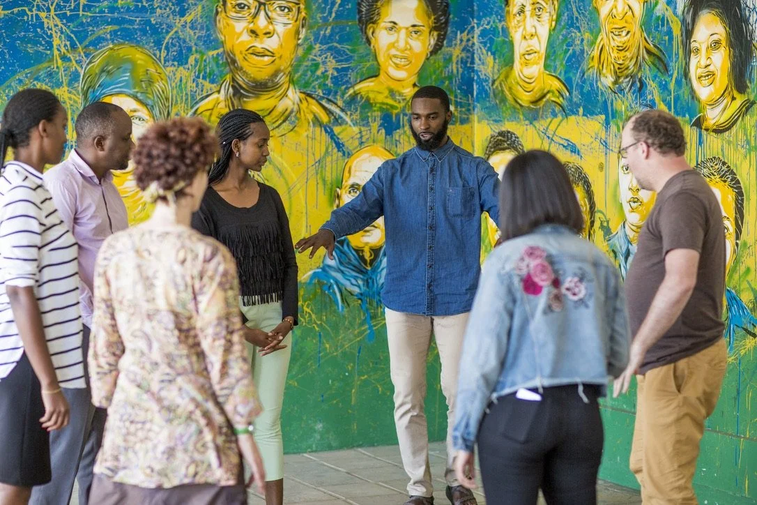 A group of diverse people standing in a semi-circle around a man who appears to be leading or instructing them, with a colorful mural of faces in the background.