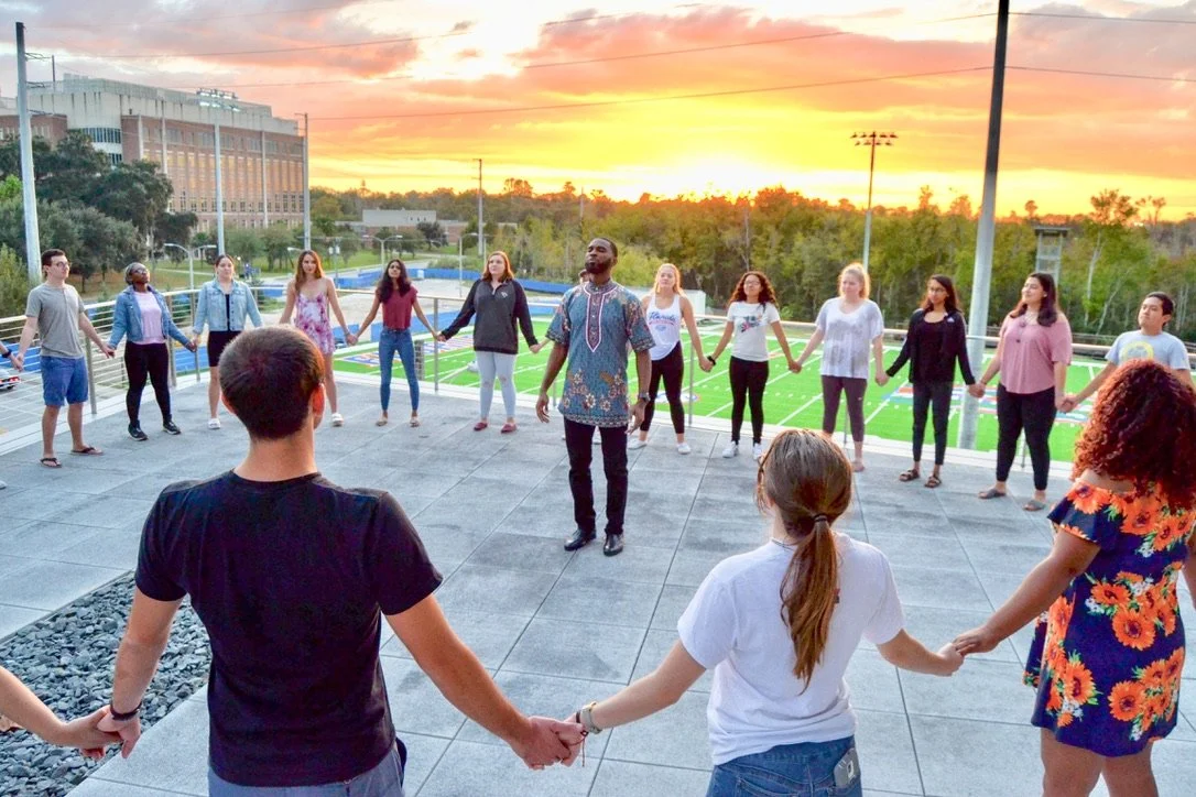 Group of diverse young people holding hands in a circle on a rooftop during sunset, near a sports field with trees in the background.