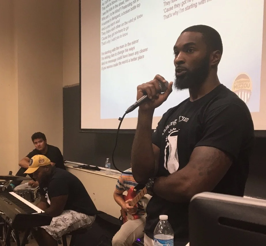 A black man holding a microphone in a classroom or lecture hall, with a large screen displaying text behind him. There are other musicians with instruments, including a keyboard, guitar, and drums, suggesting a music or performance event.