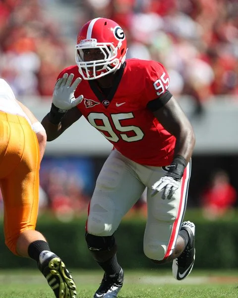 A football player from the University of Georgia wearing a red jersey with the number 95, white pants with red and black stripes, and a red helmet with a white G logo, in a defensive stance during a game.