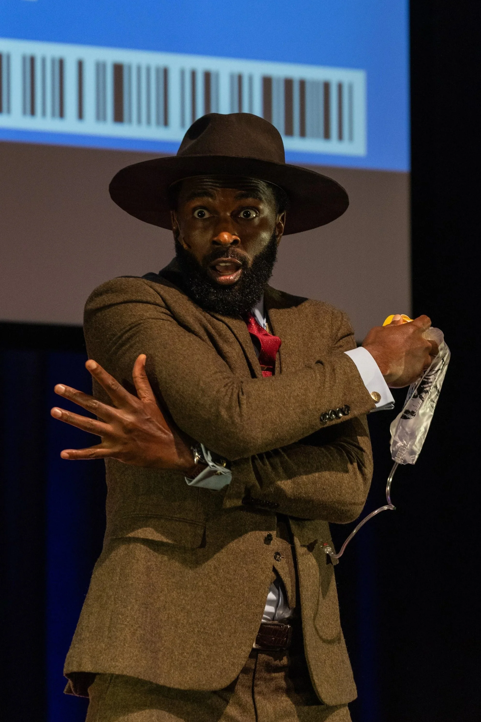 A man in a brown suit and wide-brimmed hat looks surprised, holding a wired microphone in front of a projection screen with a barcode.