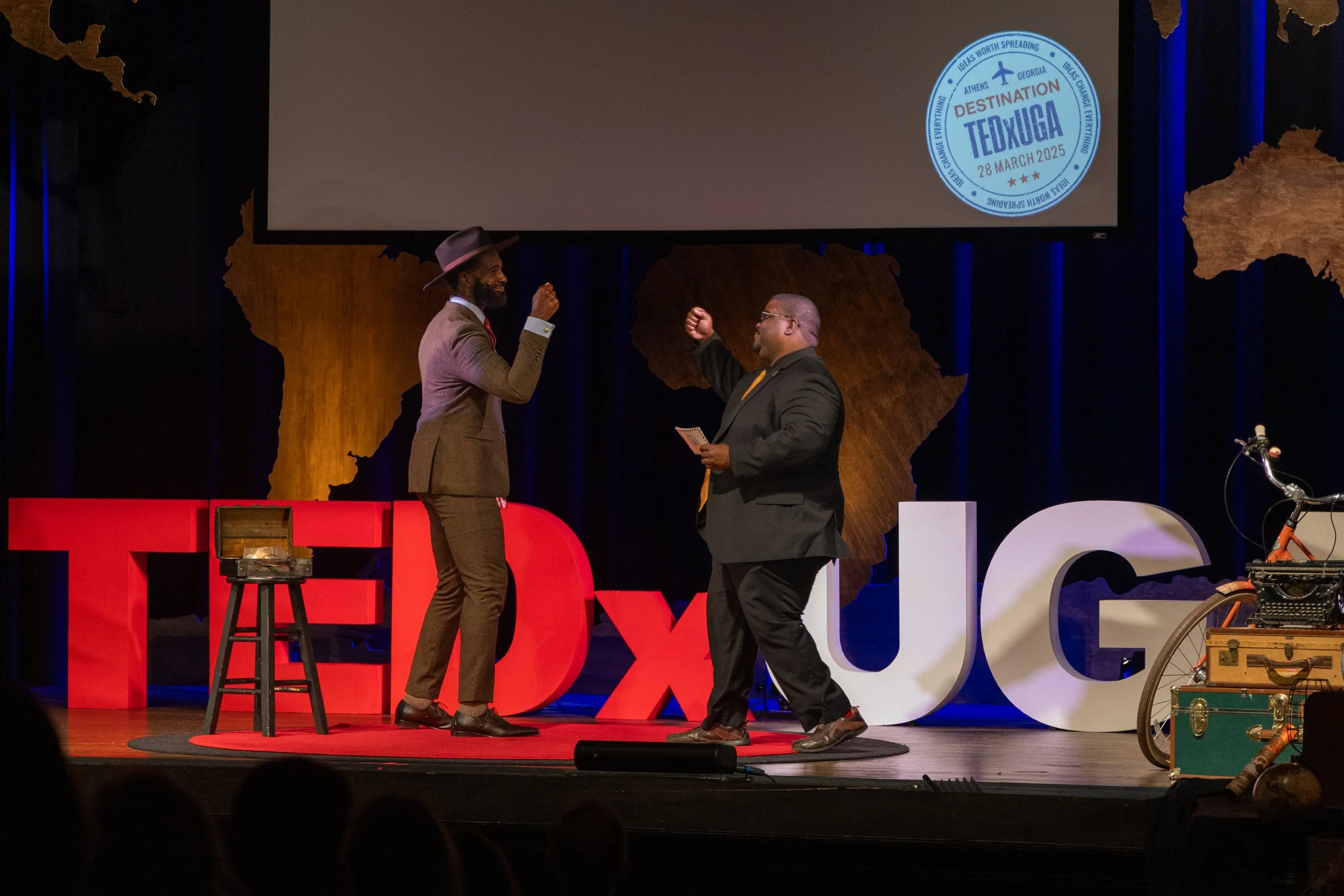 Two men on stage, one wearing a brown suit and fedora, the other in a dark suit with a yellow tie, engaging in a fist bump during a TEDx talk about TEDxUGA with a backdrop of a map of Africa and a screen displaying event details.