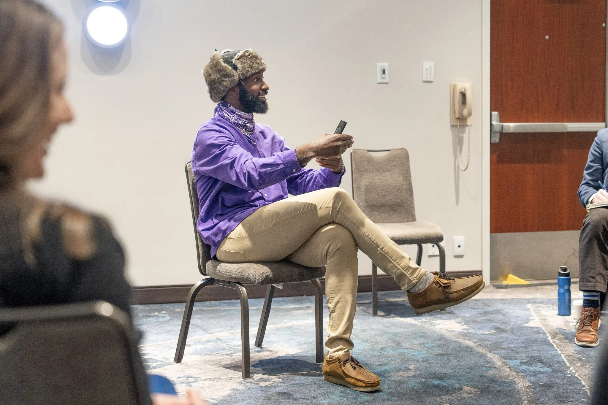 A man with a dark beard wearing a fuzzy hat, purple shirt, beige pants, and tan boots sitting on a chair and talking on a mobile phone in a conference room with other people.