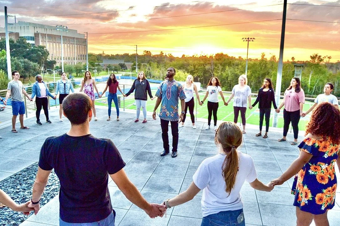Group of people holding hands in a circle on an outdoor rooftop during sunset.