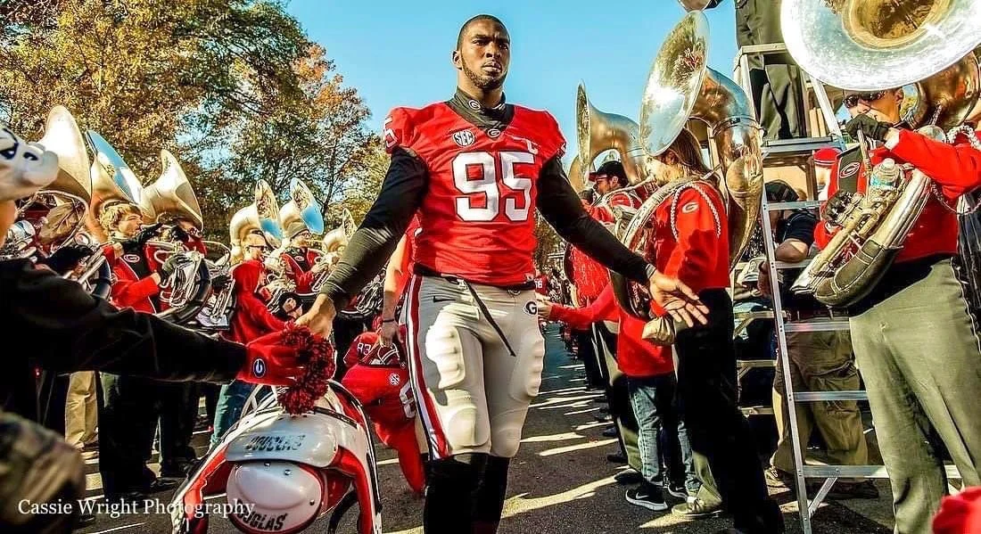A football player in a red and black uniform with the number 95, standing in the middle of a band and crowd, holding a helmet and outdoors with trees in the background.