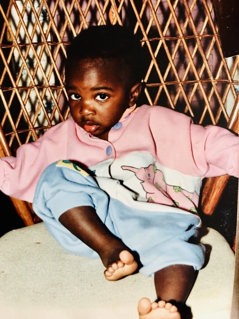 A young child with dark skin and short hair sitting in a woven rattan chair, wearing a pink top and light blue pants, with one leg crossed over the other and feet visible.