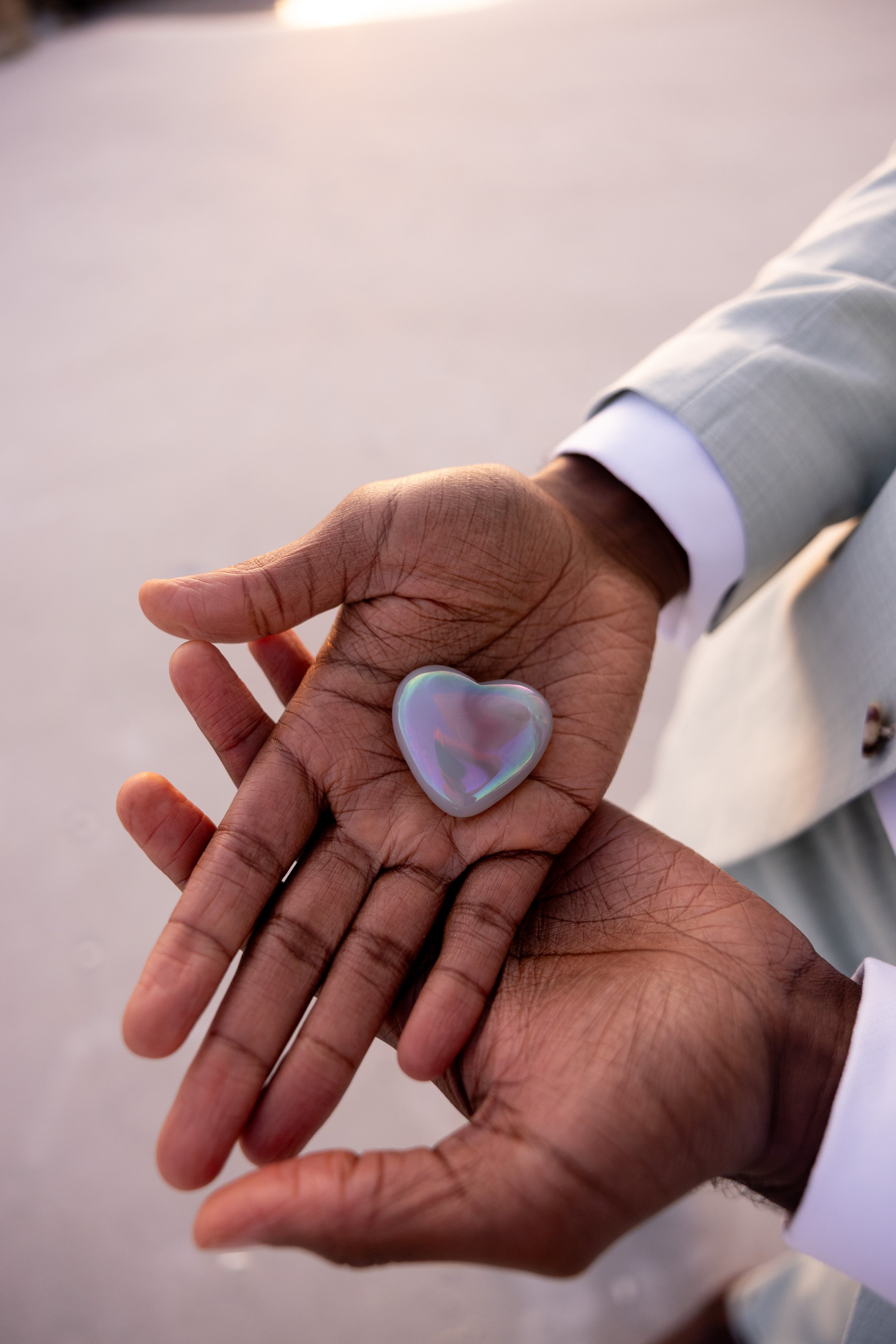 Close-up of two hands holding a pocket-sized, iridescent heart-shaped object. One hand appears darker and the other lighter, possibly indicating different skin tones. The background is plain and blurred.