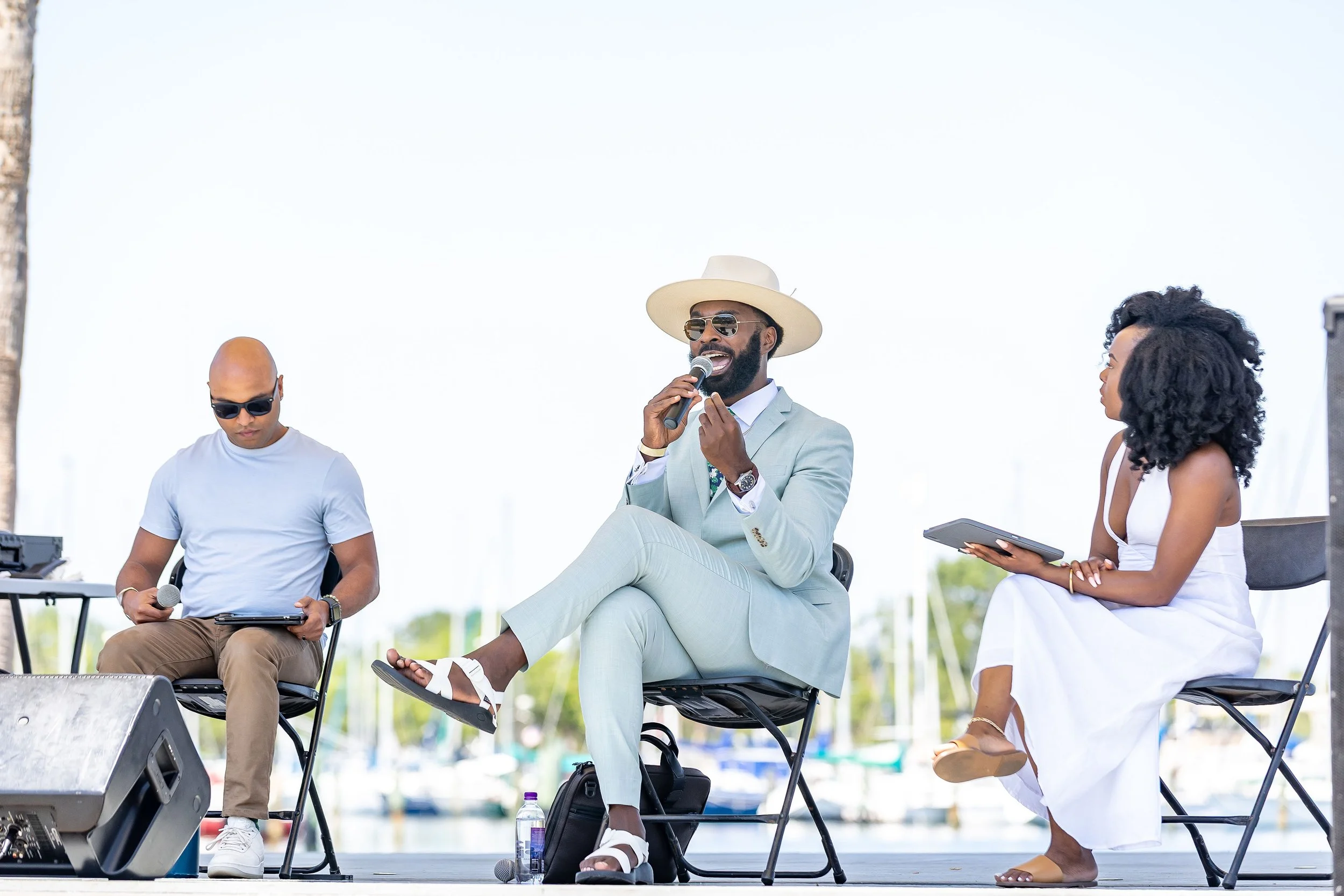 Three people sitting on a stage near a body of water, with a man in the middle speaking into a microphone. The man is wearing a light blue suit, white hat, sunglasses, and has a beard. The man to the left is wearing sunglasses, a white t-shirt, and b