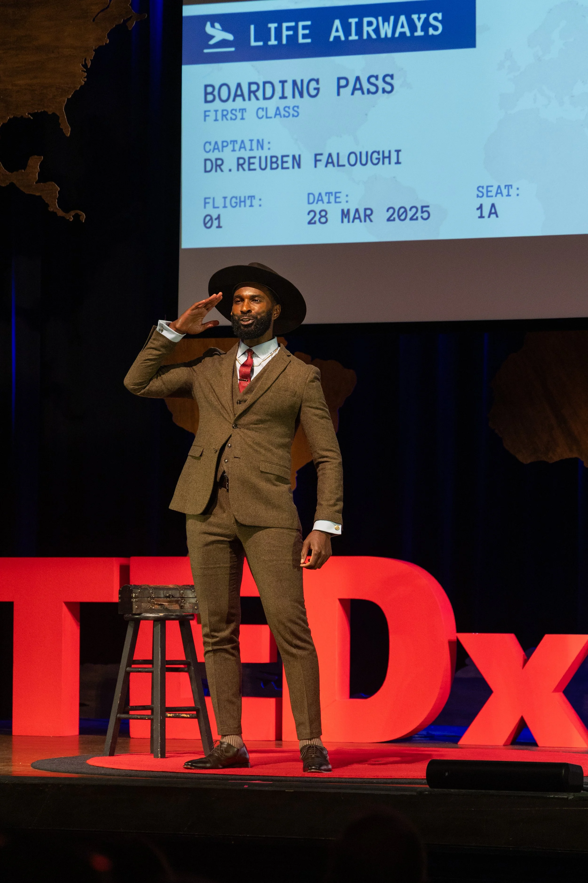 A man dressed in a brown suit and hat on a TED stage, saluting in front of a large screen displaying a boarding pass for Life Airways with details for Dr. Reuben Faloughi's flight.