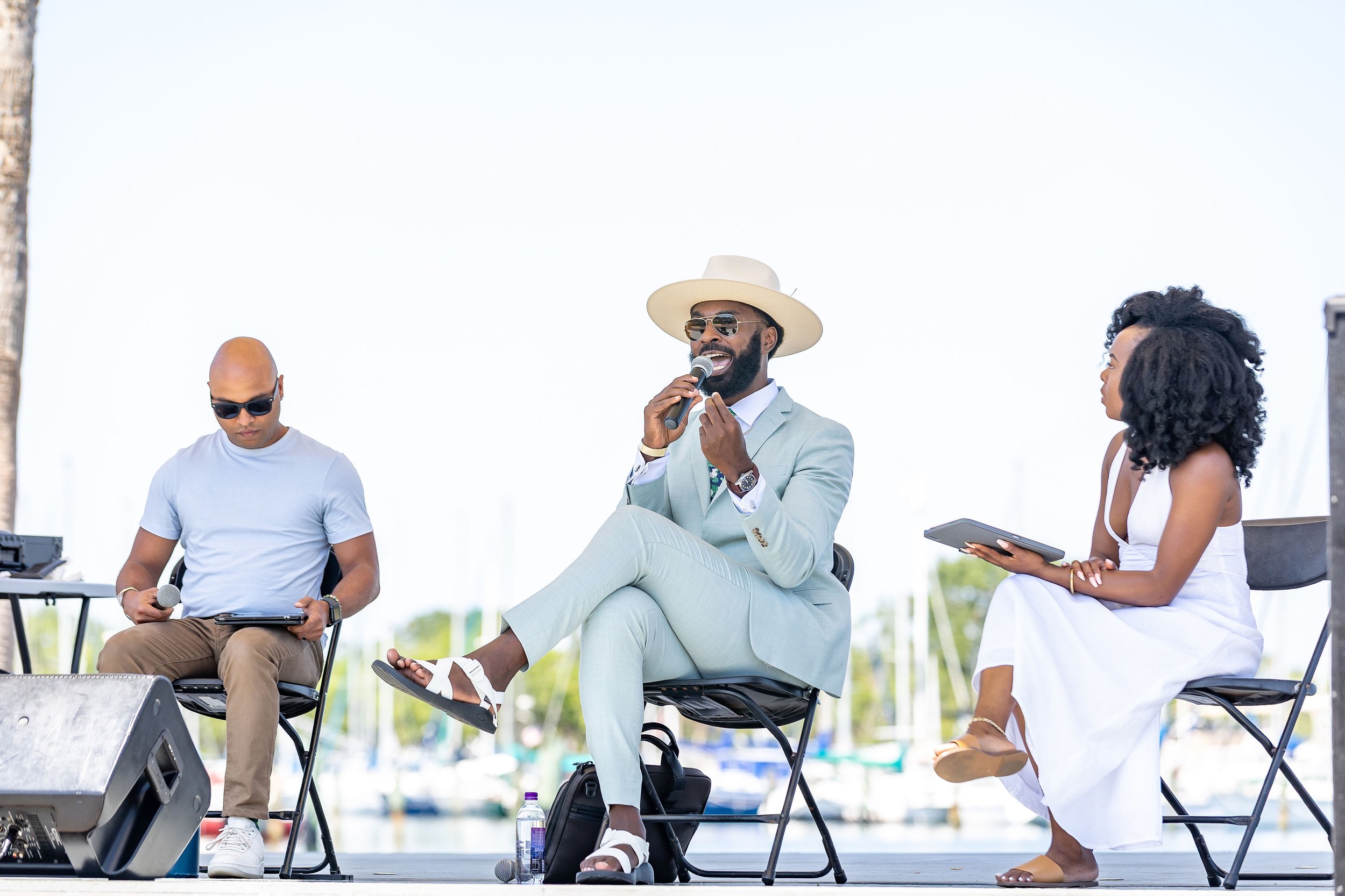 A man in a light gray suit with a white hat and sunglasses is holding a microphone and speaking, seated on a chair. To his right, a woman in a white dress with curly hair is holding a tablet, and to his left, a man in a white t-shirt with sunglasses 