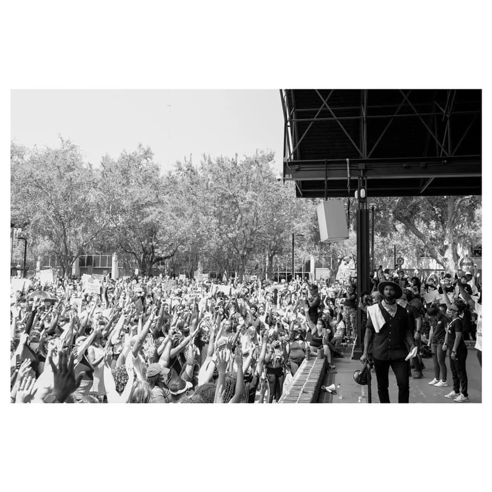 A large outdoor protest or rally with a crowd of people holding signs and raising hands, with some police present and trees in the background.