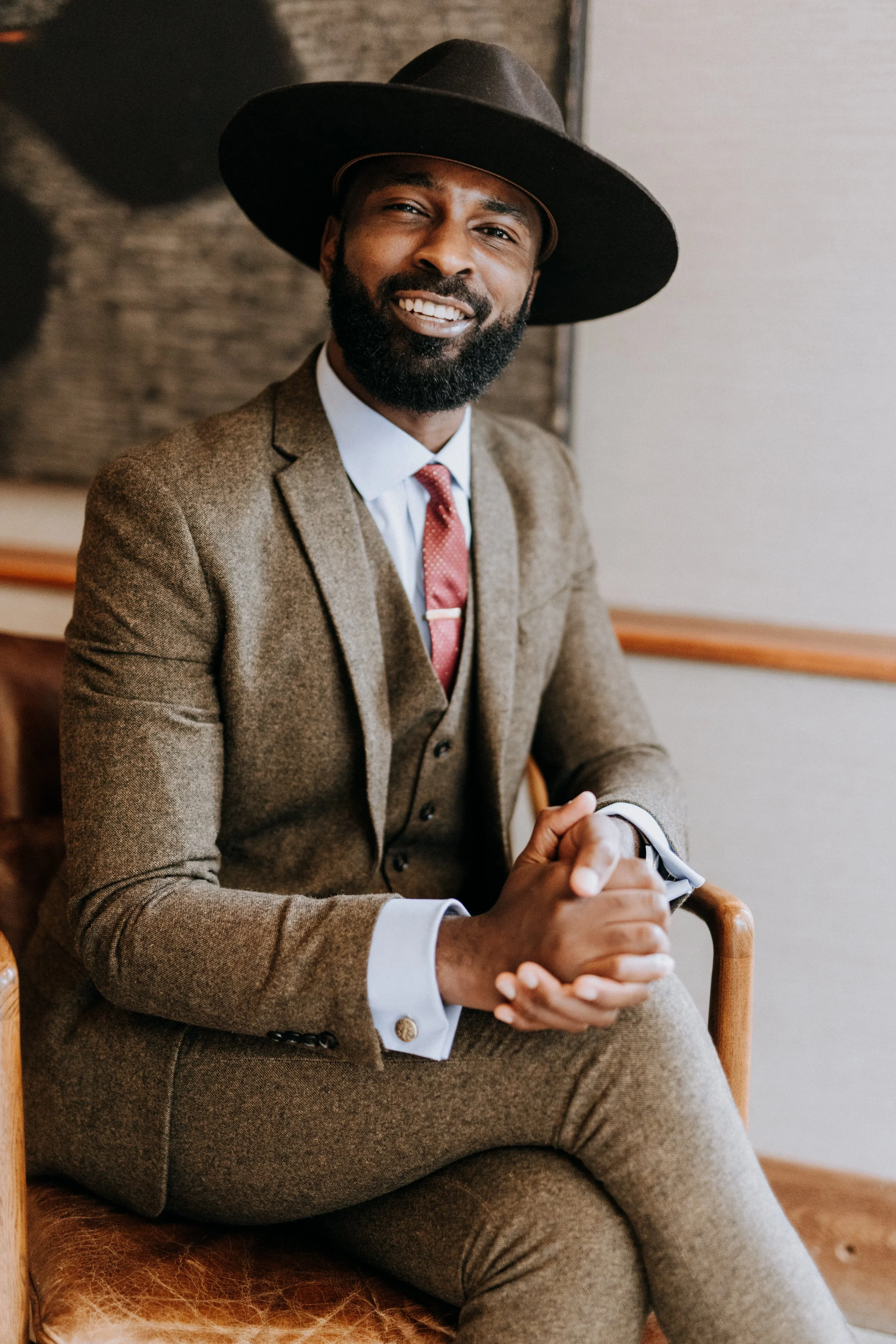 A man with a beard wearing a dark wide-brimmed hat, a brown tweed suit, a white shirt, and a red tie, sitting in a chair with his hands clasped together, smiling at the camera.