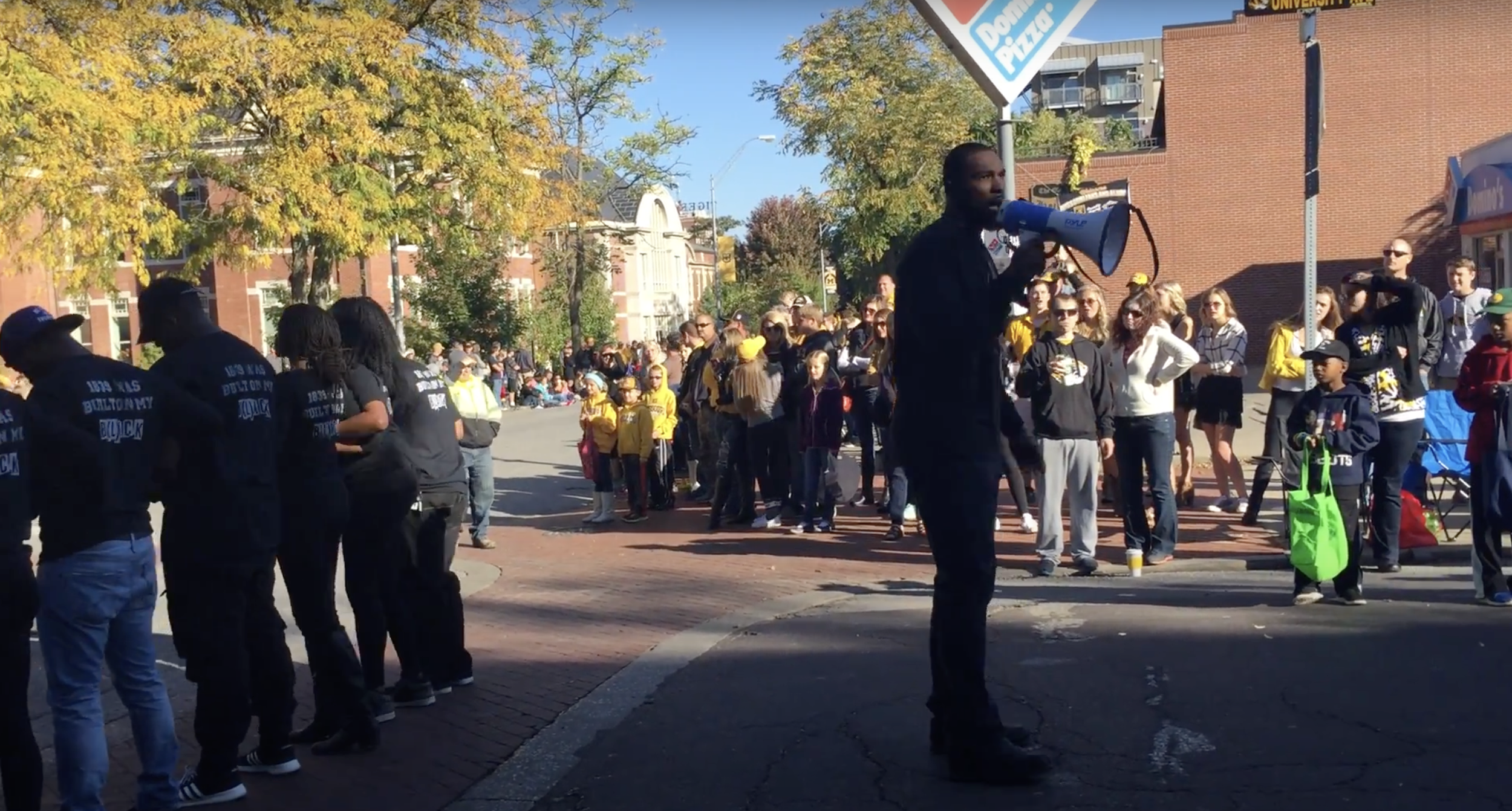 A crowd of people gathered outdoors on a city street corner, with some holding signs and wearing yellow hats, while one person with a megaphone addresses the crowd. Autumn trees with yellow leaves are visible in the background.