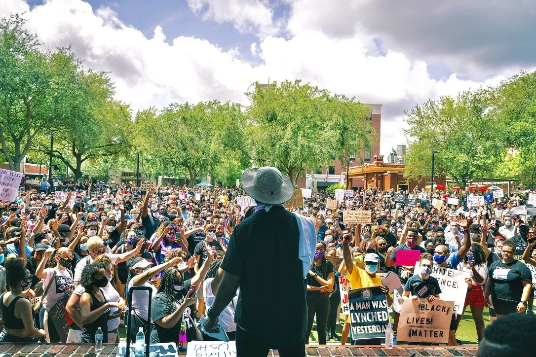 Crowd of protesters with signs, many wearing masks, gathered outdoors on a sunny day surrounded by green trees.
