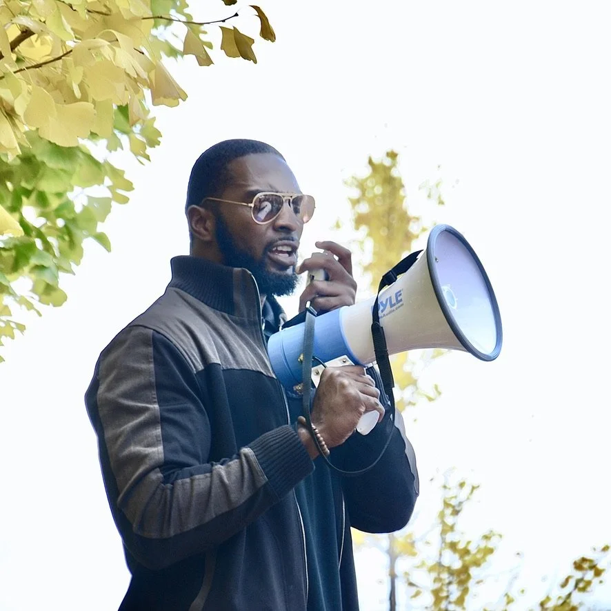 A man wearing sunglasses and a jacket holding a megaphone outdoors.