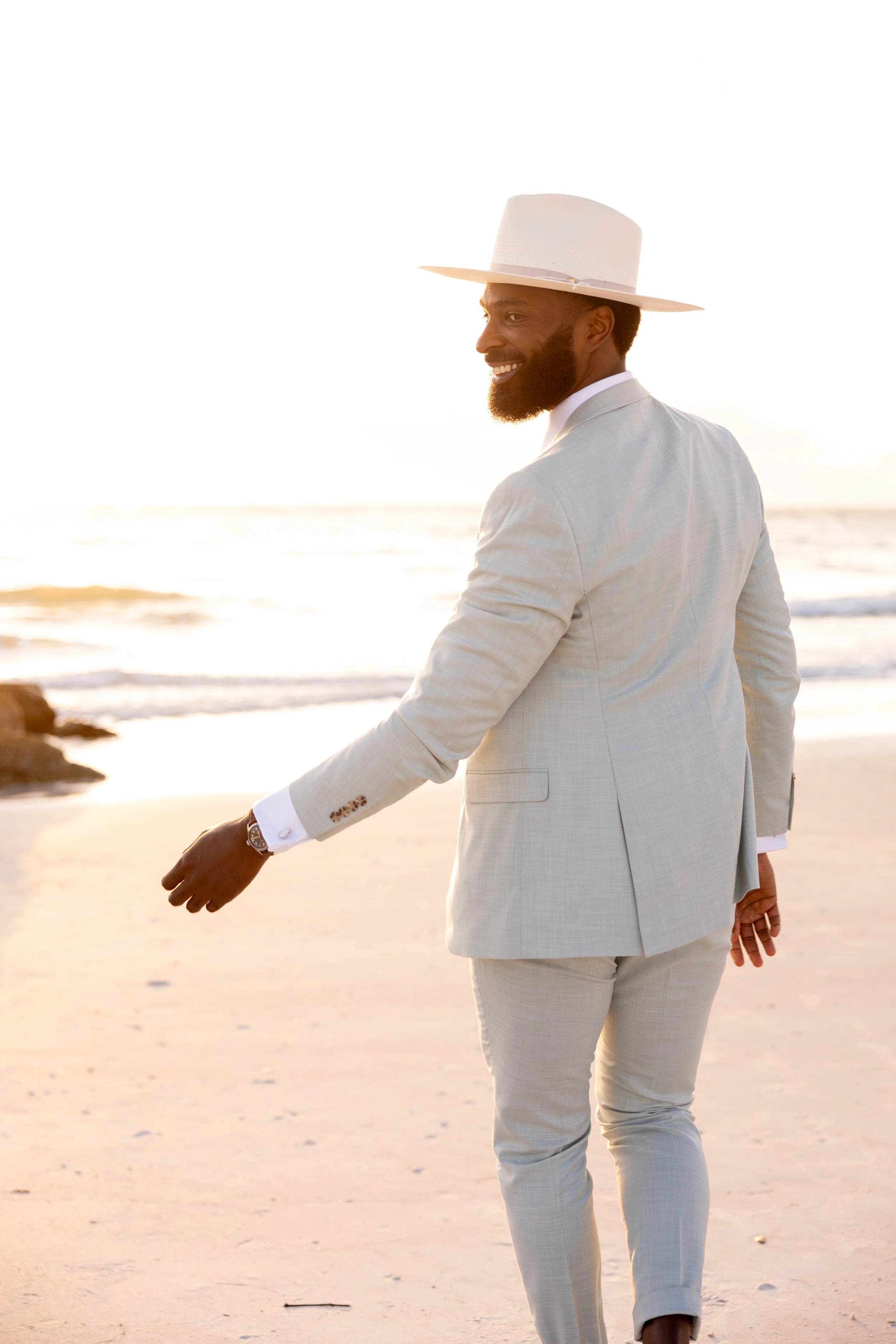 A man in a light-colored suit and a wide-brimmed hat walking on a beach at sunset, smiling and looking back.