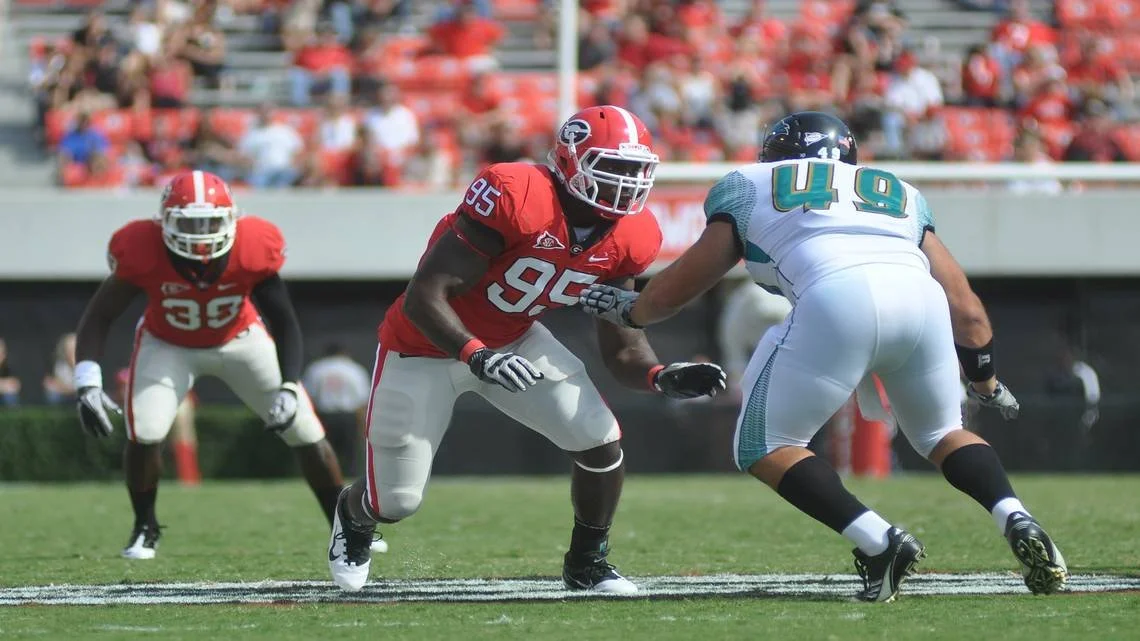 American football game with a player in red jersey number 95 blocking a player in white and teal jersey number 49, while another red-jersey player watches in the background.