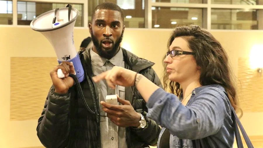 A man holding a megaphone and a woman pointing at his phone while speaking to him indoors.