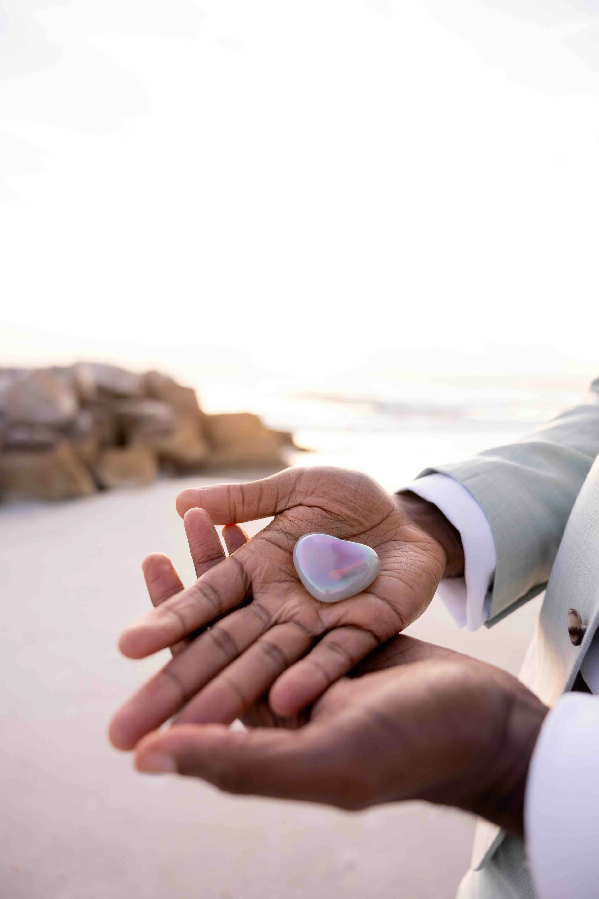 A person in a light-colored suit is holding a white, iridescent, heart-shaped object in the palm of their hand, with a beach and rocks in the background.